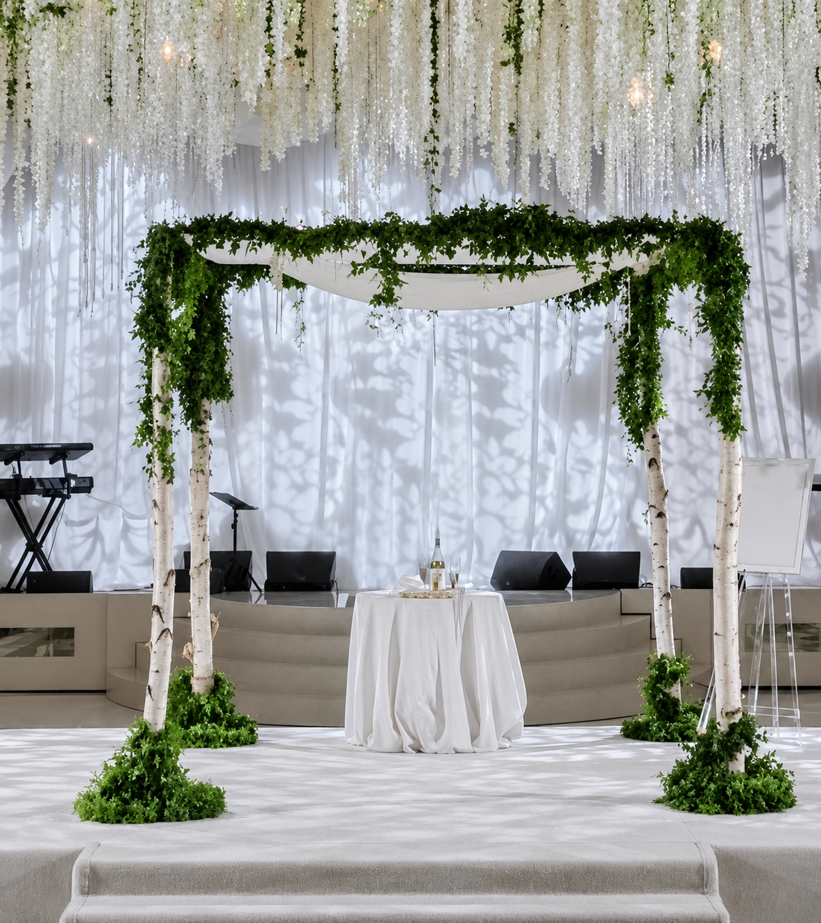 Wedding or event stage decorated with greenery and white drapery, with a table set with champagne bottles, and musical instruments on the side. BIRCH TREE CHUPAH