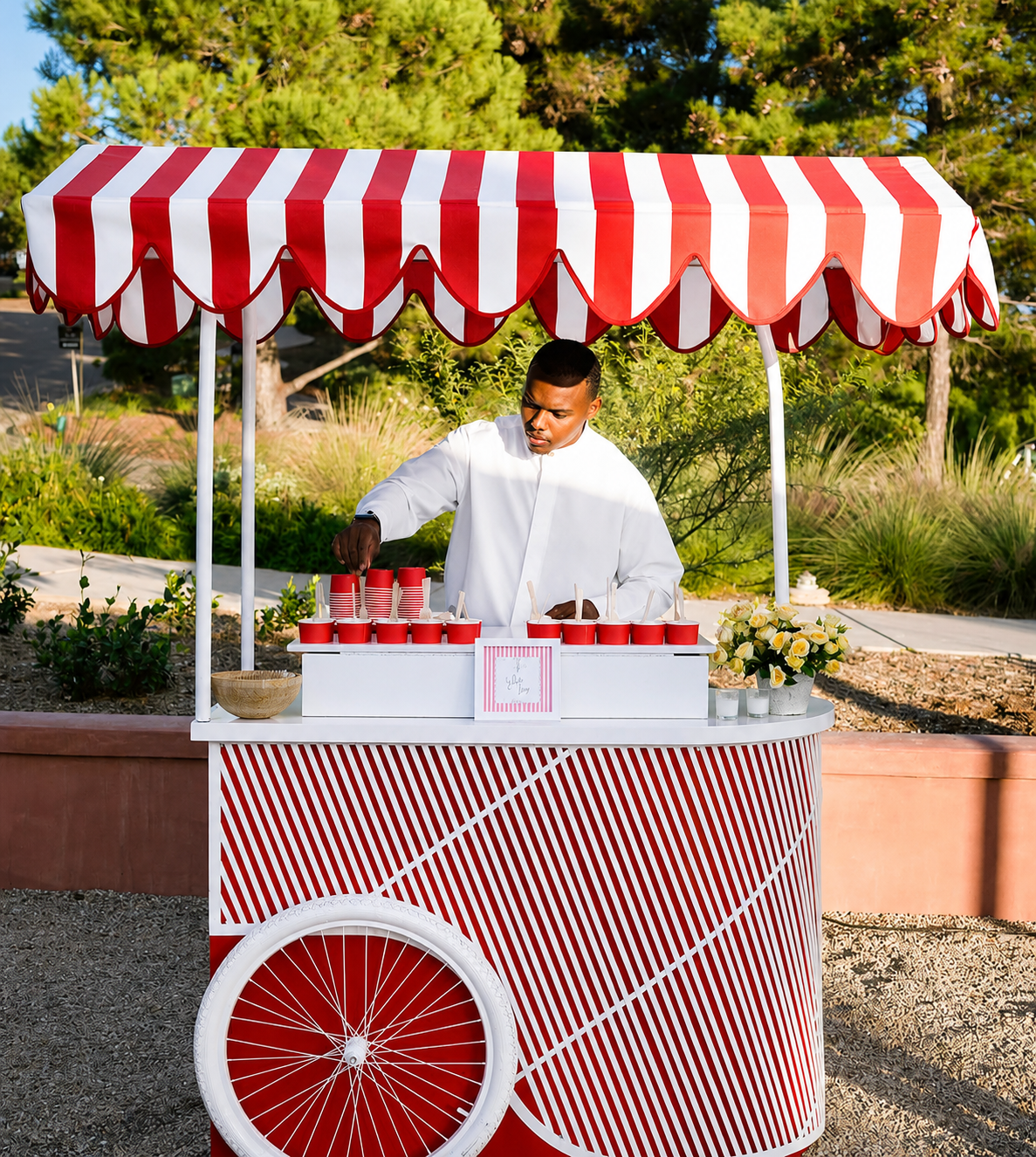 A man in a white shirt working at a red and white striped ice cream cart with a matching striped umbrella outdoors surrounded by greenery.