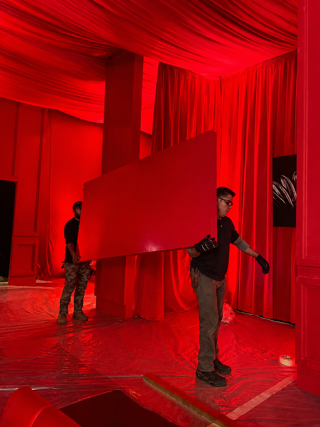 Two workers setting up a red stage with draped curtains, one holding a large red panel, in an indoor event space.