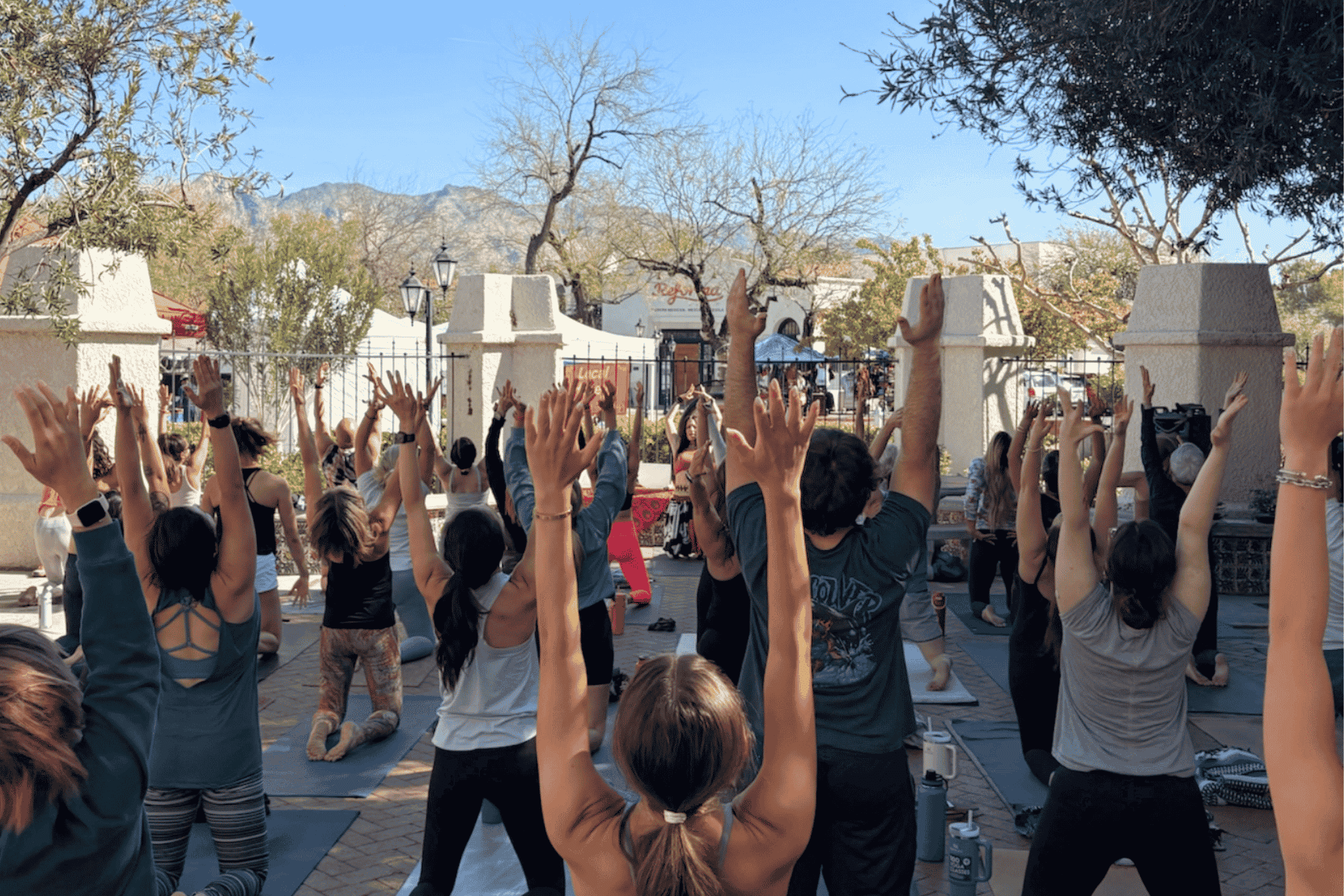 A large group of people practicing yoga outdoors in the courtyard of yoga pod tucson, mountains in the background, and clear blue sky.