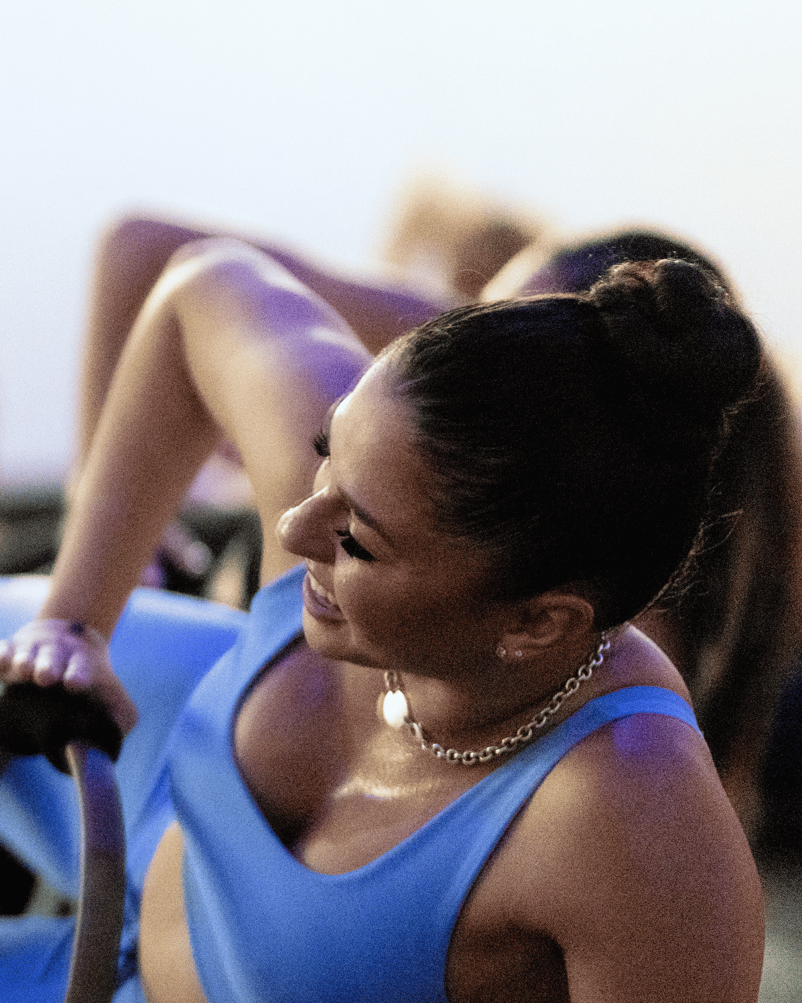 A woman with dark hair styled in a bun wearing a blue sports top, gold jewelry, and smiling, with other women in the background, practicing barre at yoga pod tucson.