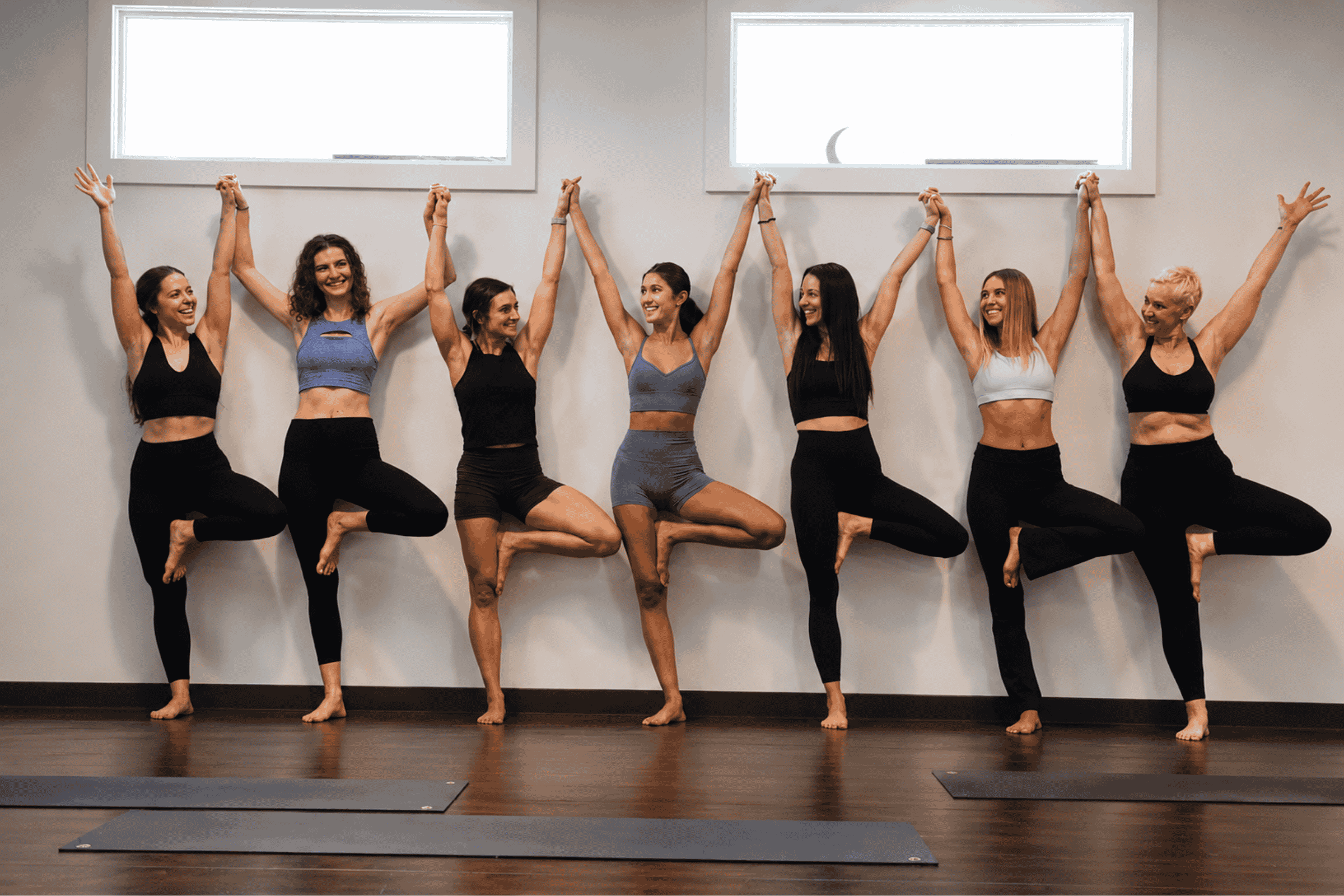 Smiling women in tree pose holding hands overhead along a studio wall at Yoga Pod Tucson