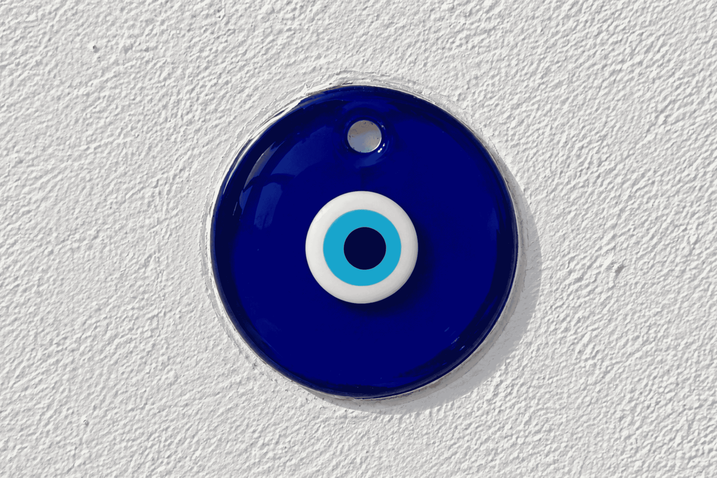 Blue glass evil eye charm with a white and light blue center against a white textured wall