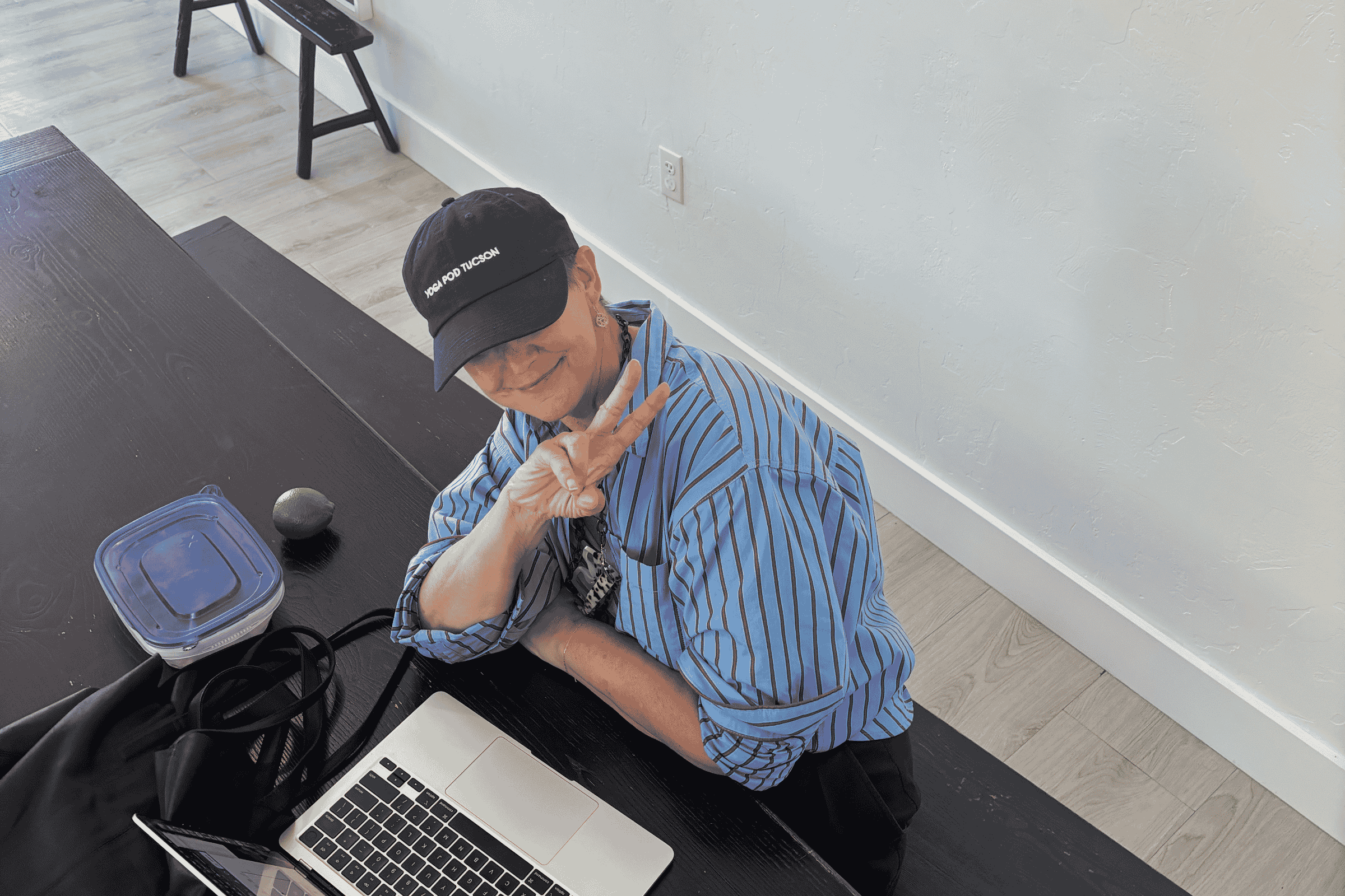 Person in a Yoga Pod Tucson cap and striped shirt flashing a peace sign from a table with a laptop
