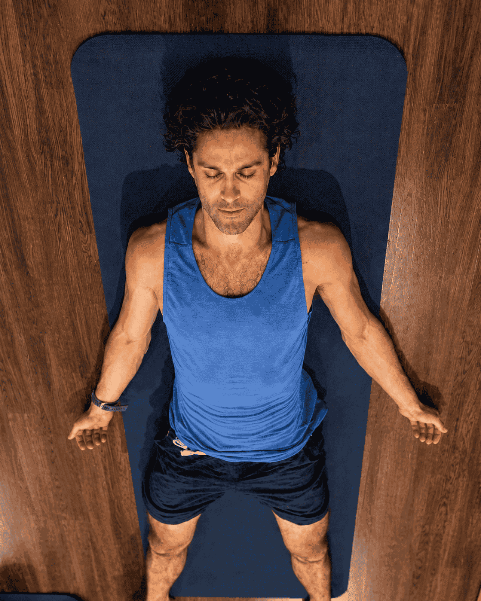 A man lying on his back on a yoga mat with eyes closed, wearing a blue tank top and black shorts, surrounded by wooden floors.