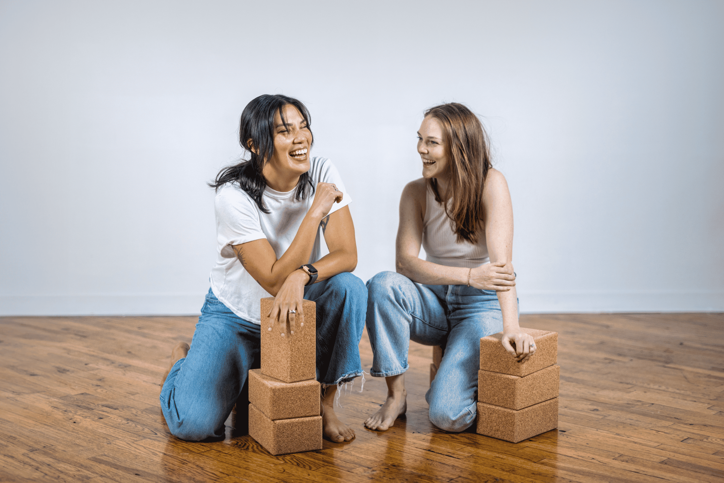 Two women sitting on wooden floor with yoga blocks and bricks, laughing and looking at each other.