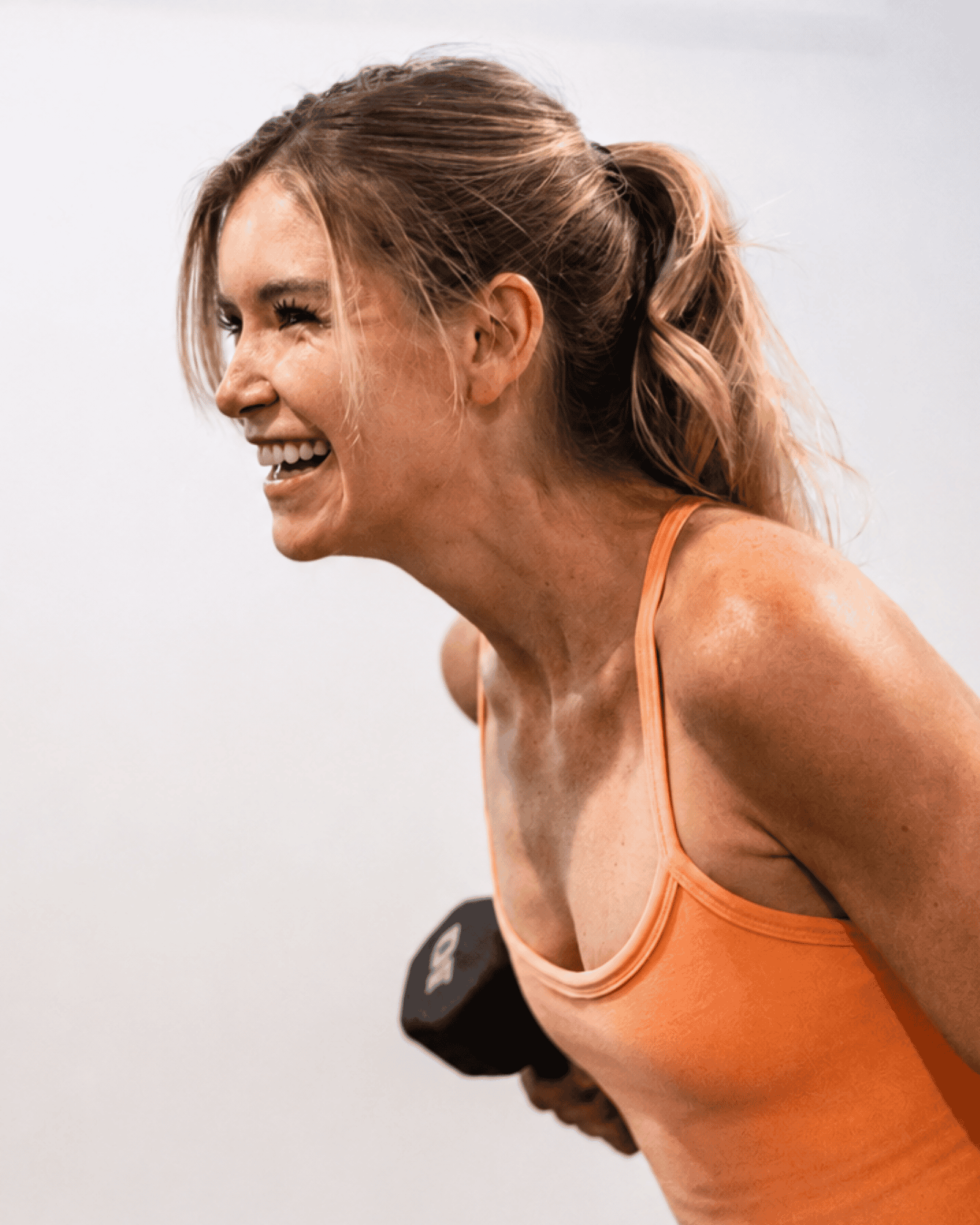 Close-up of a woman exercising, smiling, wearing an orange tank top, with a white wall background at yoga pod tucson.