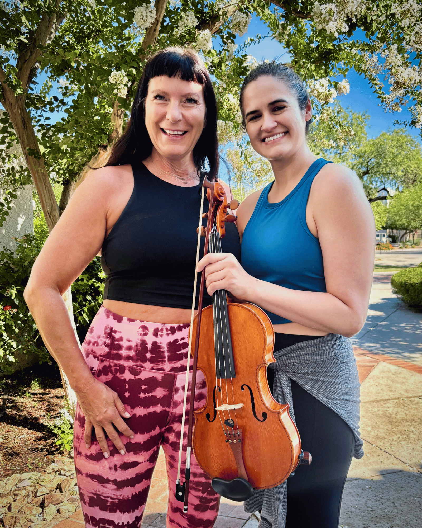Two women outdoors, one holding a violin, smiling, with trees and blue sky in the background.