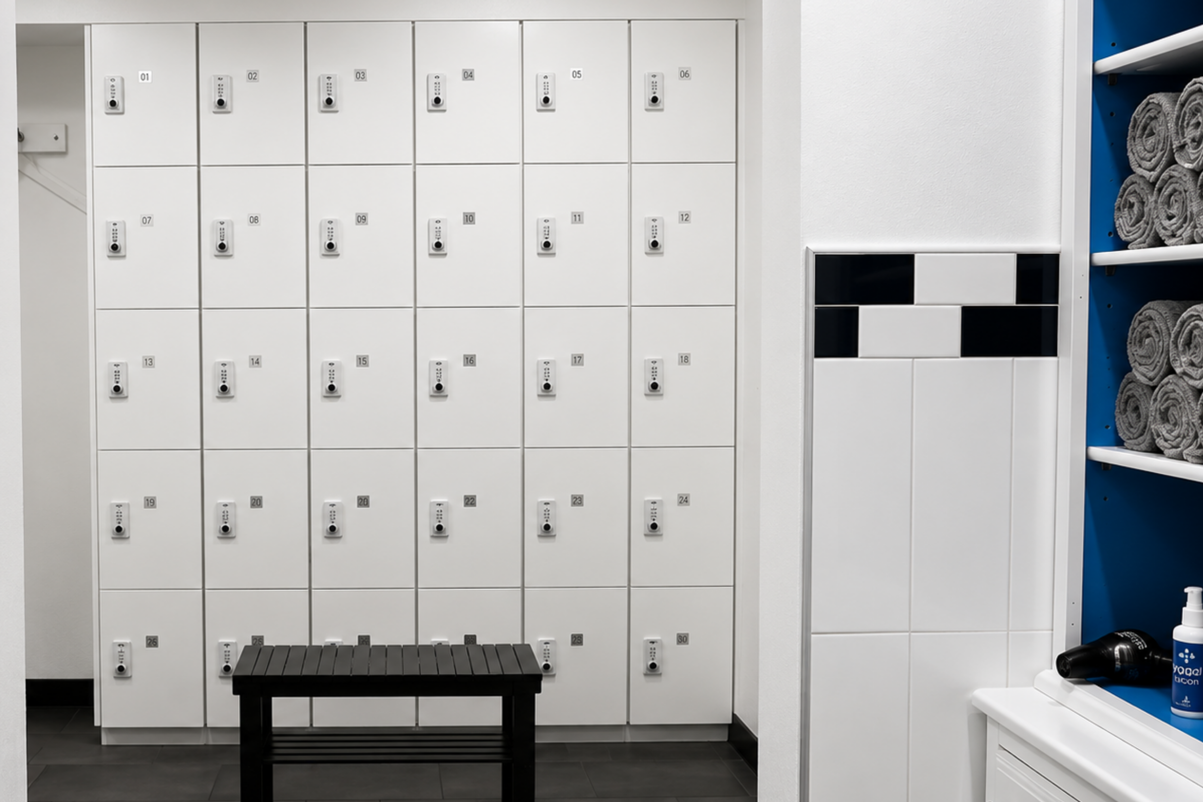 Locker room at yoga pod tucson.  Row of white lockers with combination locks, a black bench in front, and a shelf with rolled towels on the right side.