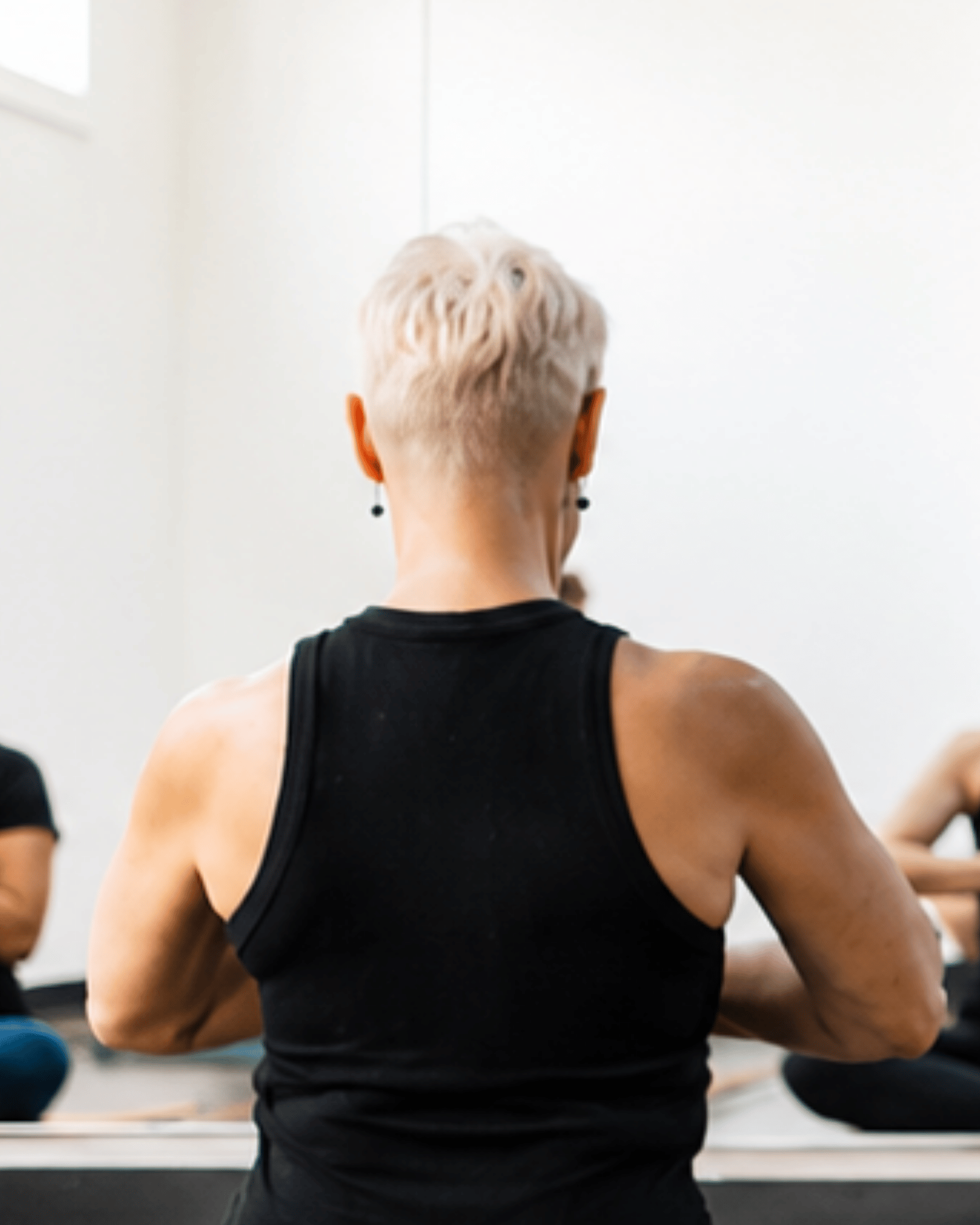 Back of a woman with bleach blonde hair practicing yoga at yoga pod tucson. She is wearing a black tank top and earrings, with her arms bent at the elbows.
