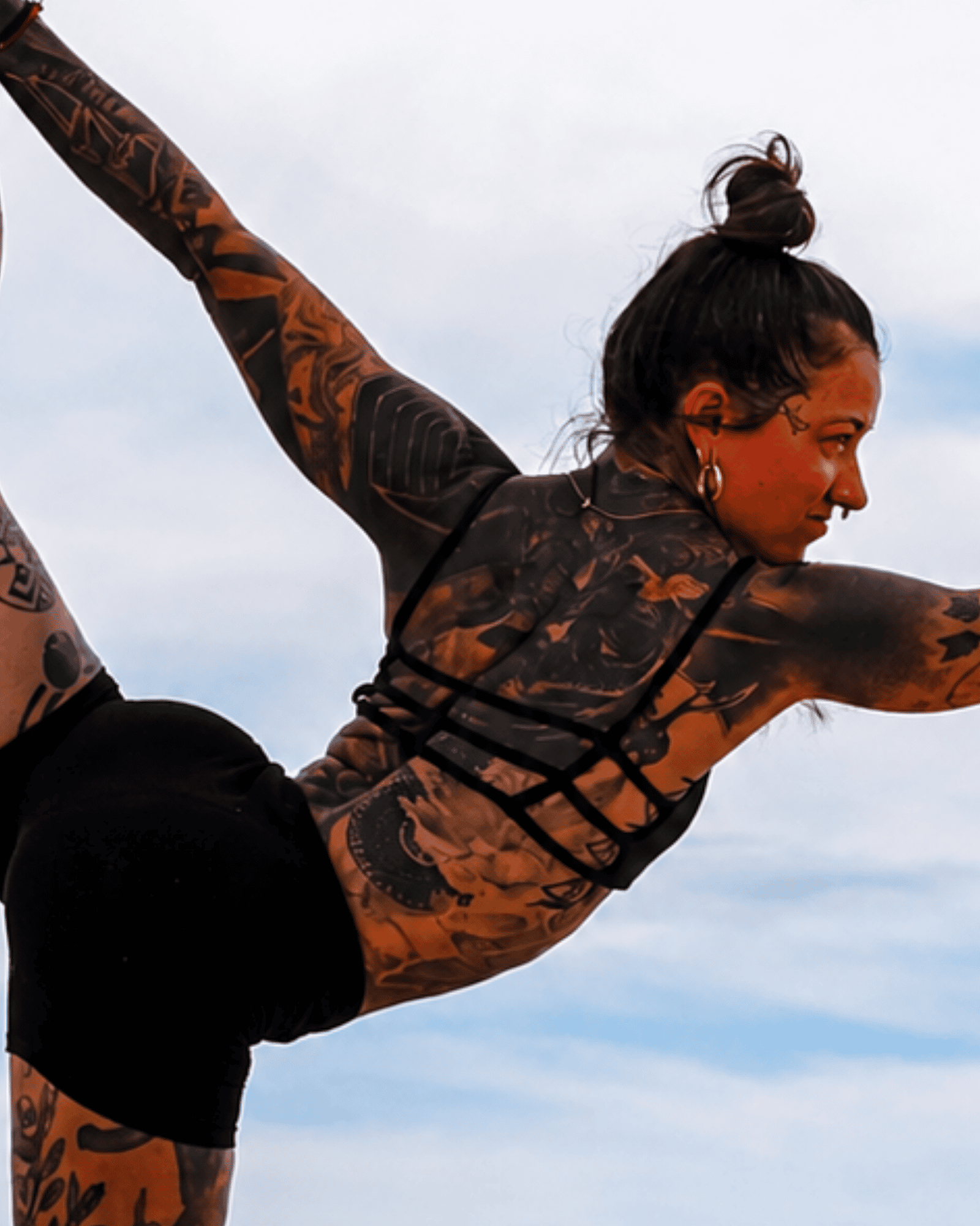 Close-up of a woman with tattoos on her arm, dark hair tied in a bun, and wearing earrings, against a cloudy sky, in a yoga or stretching pose.