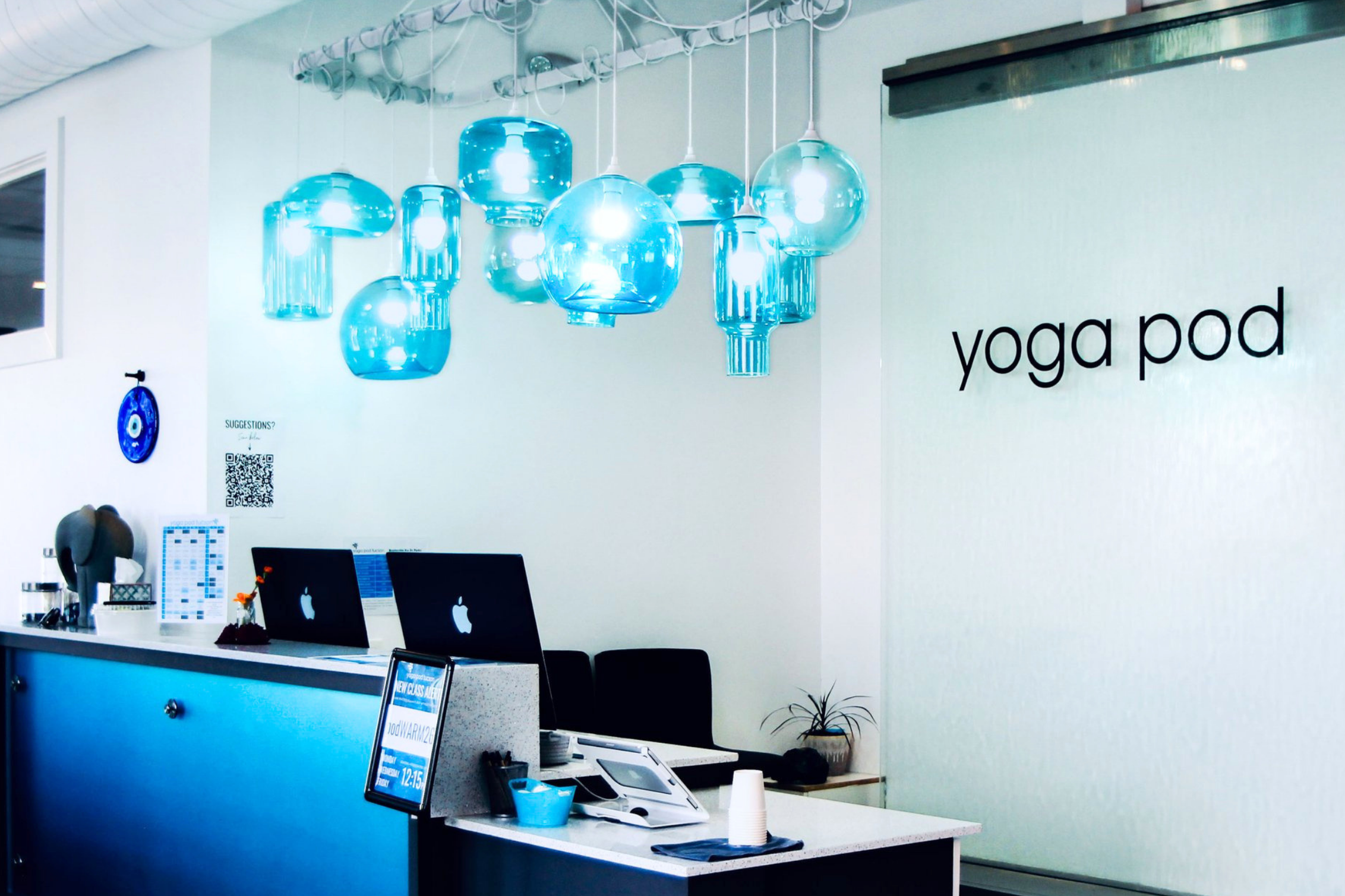 Reception desk with Apple logo computers, a QR code, digital displays, and hanging blue glass pendant lights in yoga pod tucson's modern space.