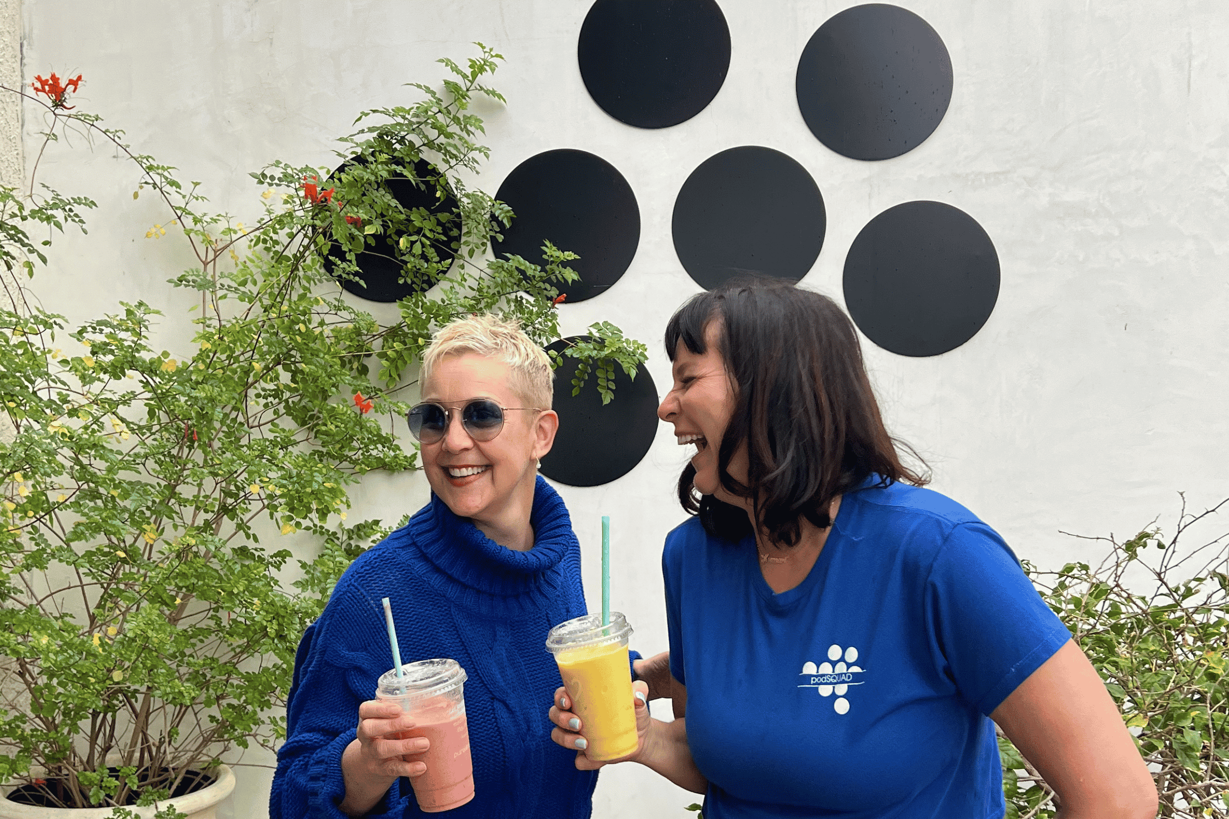 Two women enjoying smoothies outdoors, standing next to leafy green plants against a white wall decorated with black circular art pieces.