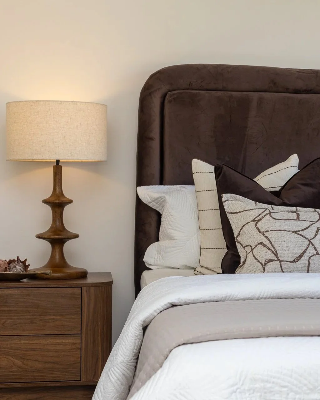 A bedroom corner with a brown upholstered headboard, pillows, and a wooden nightstand with a beige lamp, seashells, and decorative items.