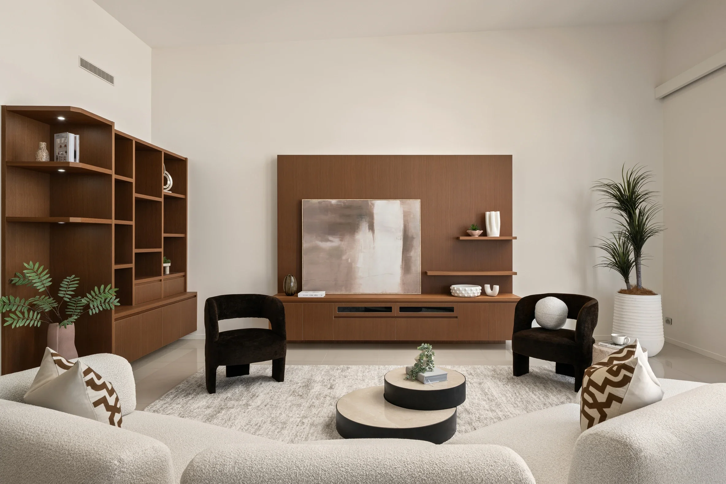 Modern living room with beige and brown furniture, including a coffee table, armchairs, and a sectional sofa, decorated with pillows and plants, with a large wooden shelving unit and a wall-mounted entertainment center.