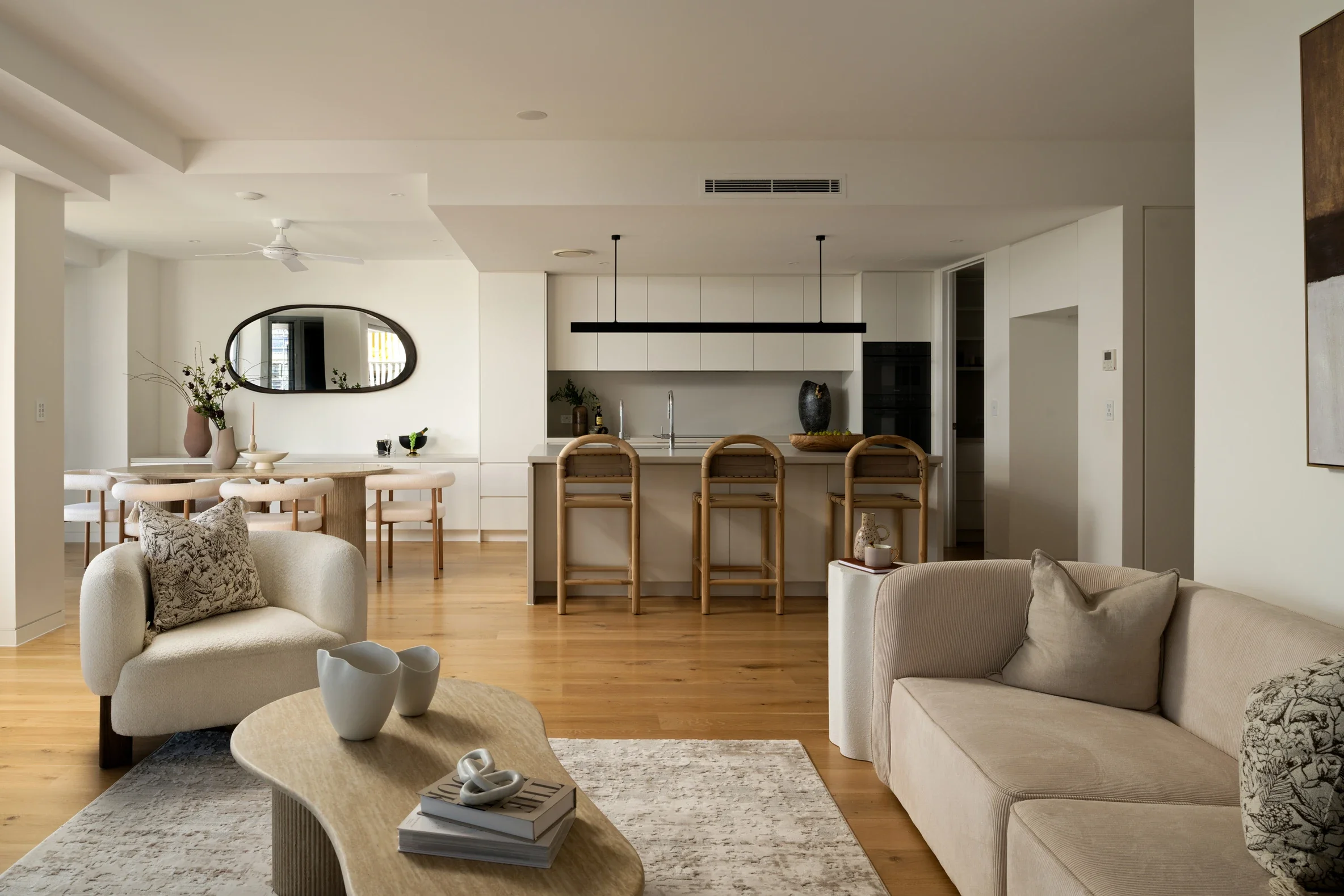 Open-concept living room and kitchen with white walls and wooden floors, featuring a sofa with pillows, a coffee table with decorative objects, a dining table with chairs, and modern kitchen cabinets with a kitchen island.