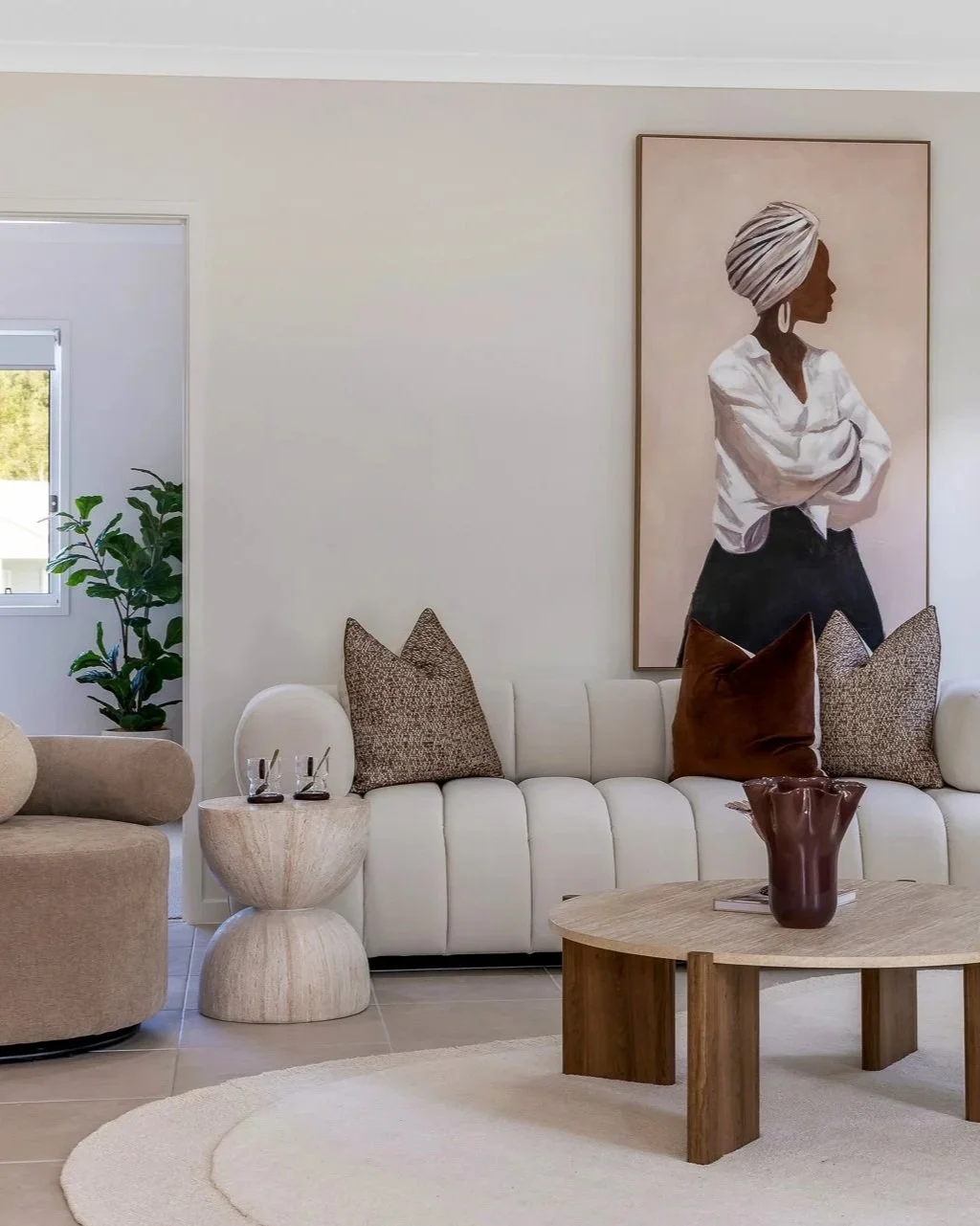 Living room with a white sofa adorned with brown and patterned throw pillows, a wooden coffee table with a brown vase, a beige armchair, a small side table with two glasses, and a large portrait of a woman with a turban hanging on the wall. A leafy green plant is visible through a nearby window.