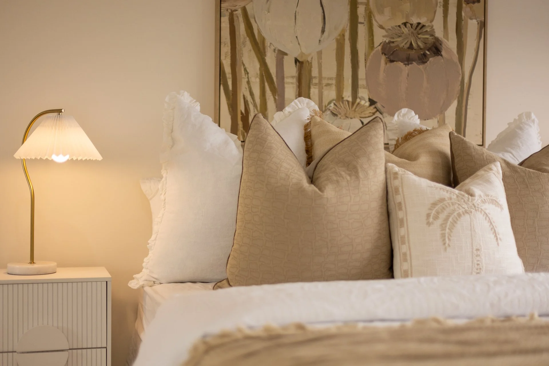 A bedroom corner featuring a tall floor lamp with a cream-colored shade, a white nightstand with vertical grooves, and a bed with multiple beige and cream pillows, a bedspread, and a large floral painting above the bed.