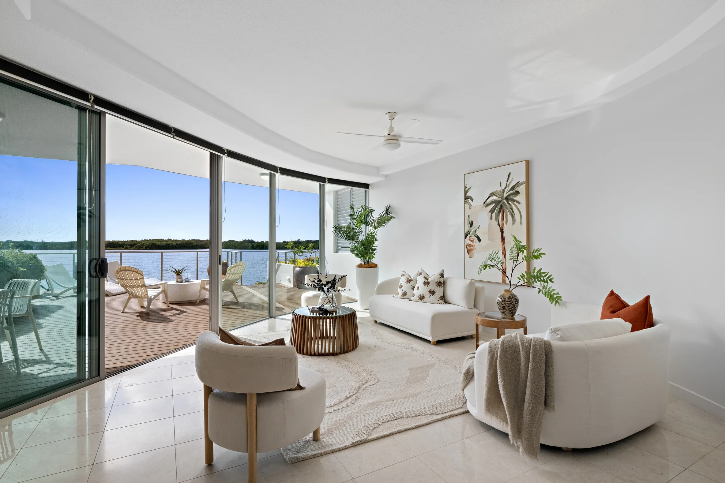 Living room with white sofa, armchair, potted plants, wall art, and large glass sliding doors opening to balcony with outdoor seating overlooking water.