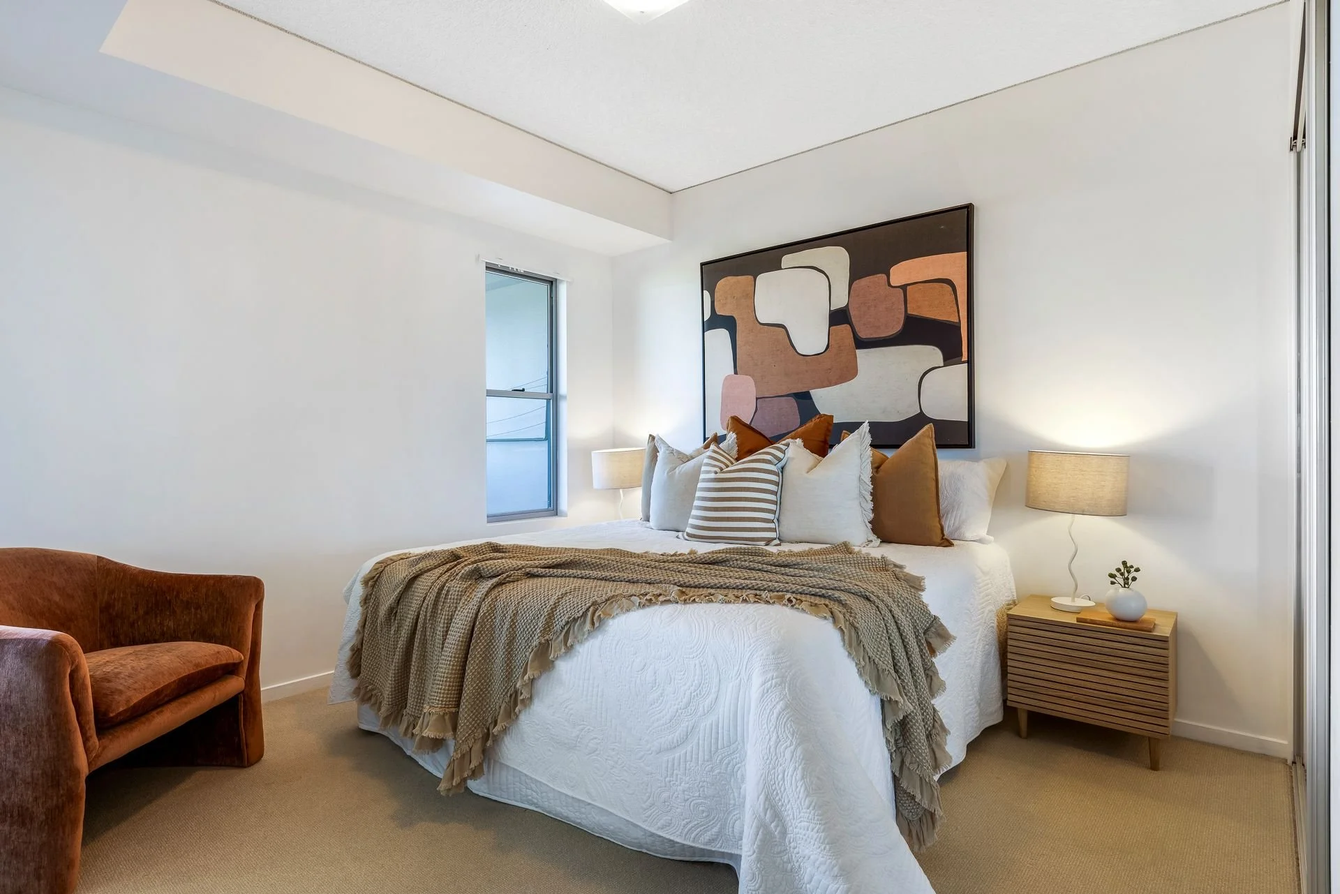 A modern bedroom with a white bed, patterned pillows, beige blanket, two bedside lamps, a small plant, a large abstract wall art, a window, and a beige carpet.