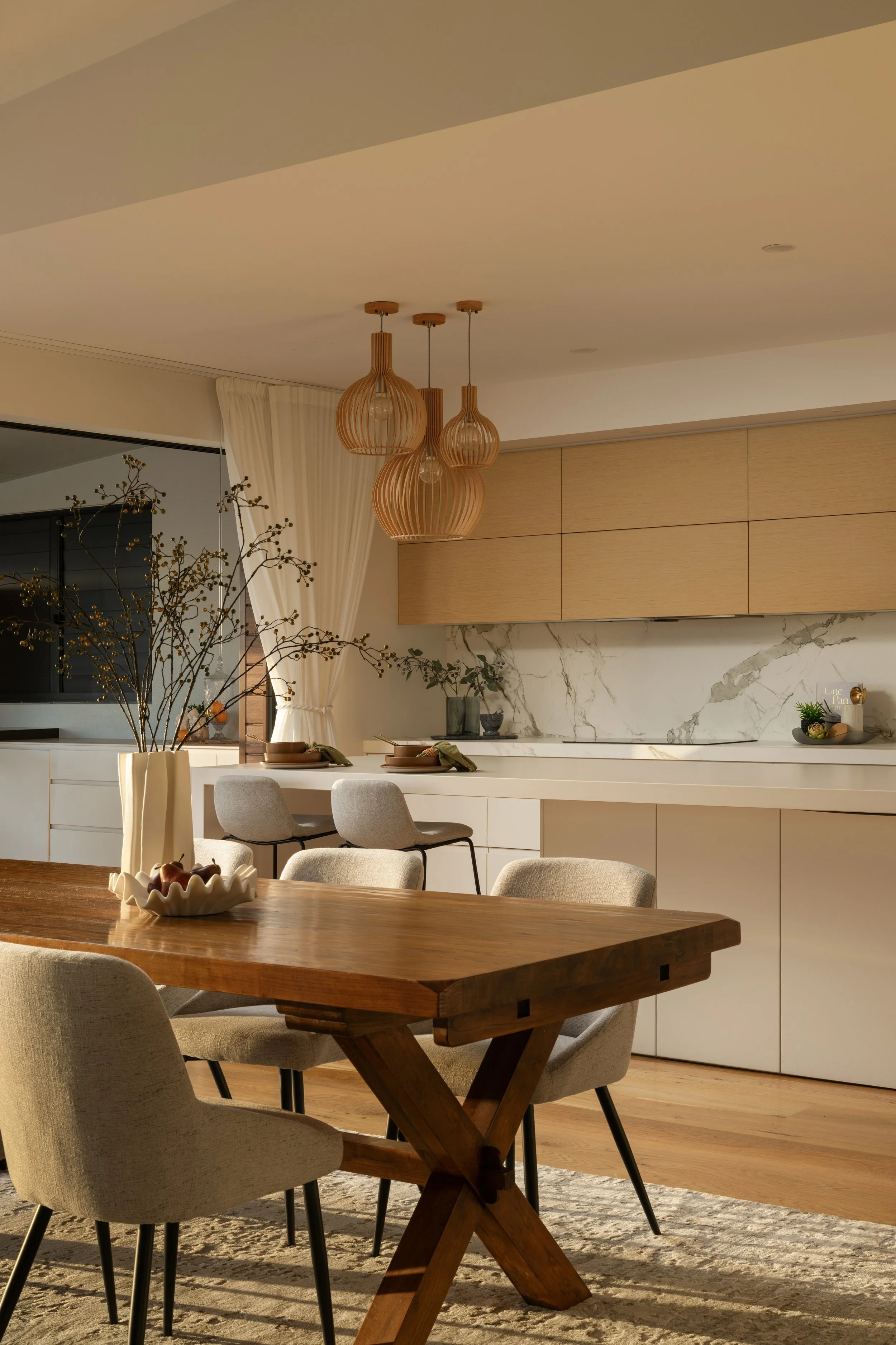 Modern dining area with a wooden table, beige upholstered chairs, decorative branches in a cream vase, pendant lights, white cabinetry, marble backsplash, and a textured rug.