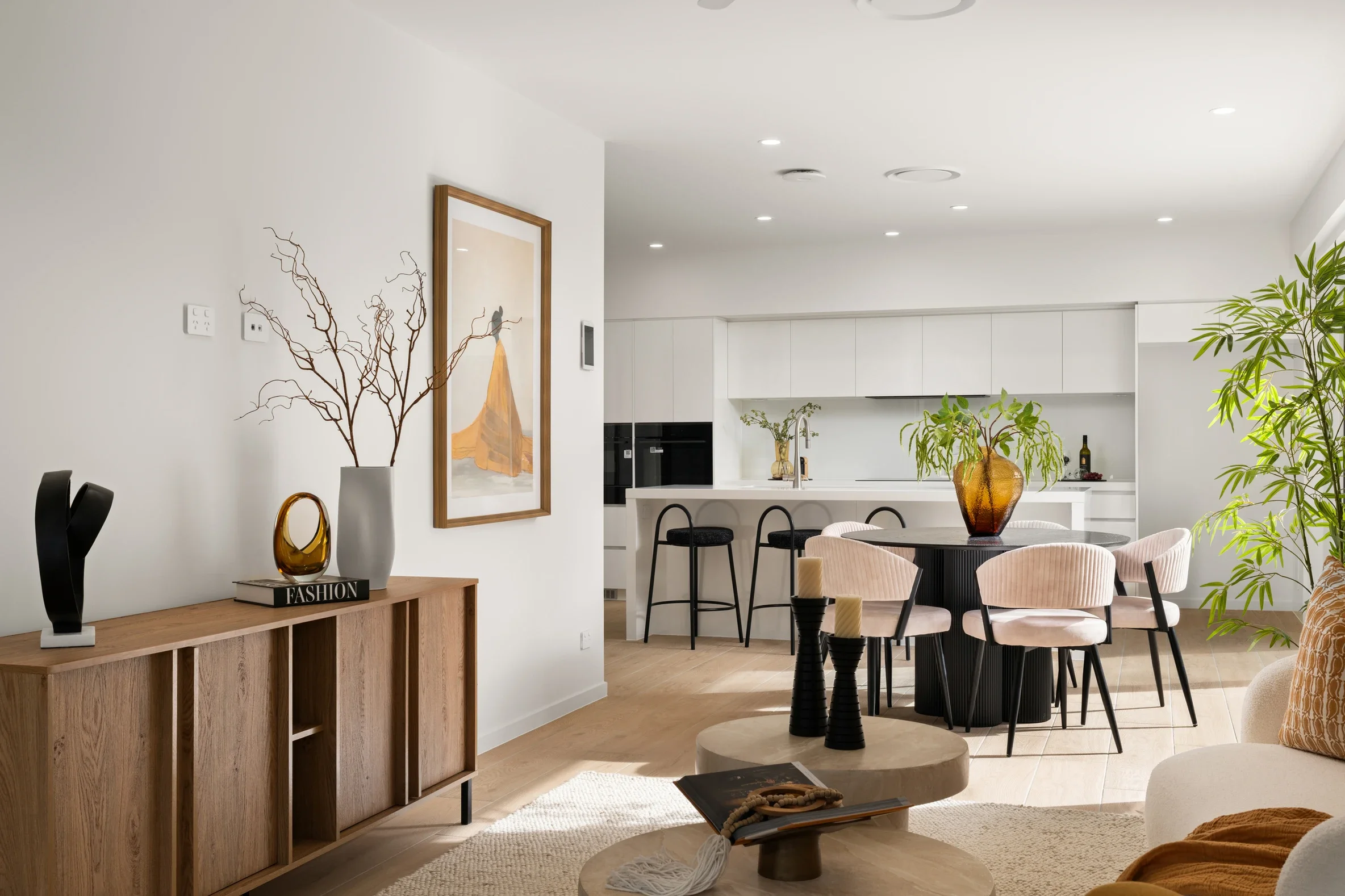 A modern open-concept living space with a dining area and kitchen. The room features white walls, light wood flooring, and contemporary decor including a round dining table with beige upholstered chairs, a wooden sideboard with decorative items, and green plants. The kitchen has white cabinetry, a white countertop, and a few visible appliances. Pendant lights are mounted on the ceiling, and the room is well-lit with natural light.