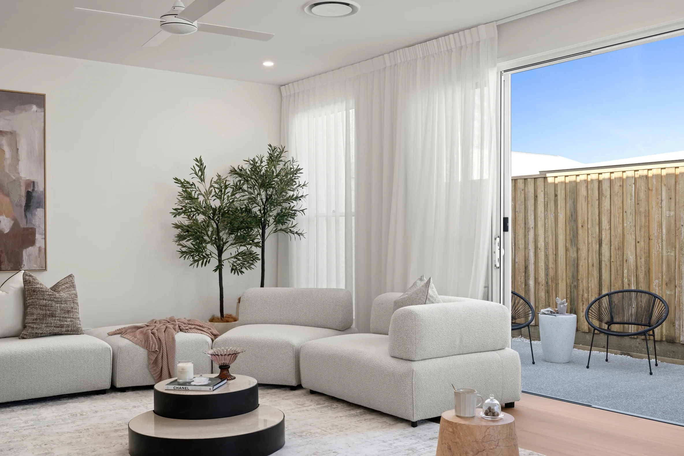 Living room with white sofa, several pillows, wood side table, potted plant, large glass door with white curtains, and outdoor seating with wooden fence and blue sky visible.