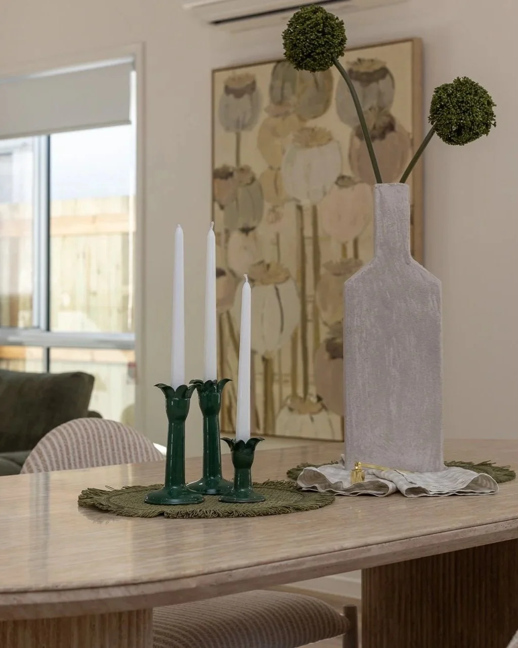 A dining table with a green candle holder with three white candles, a tall beige textured vase with two green pom-pom flowers, and a cloth napkin with a gold ring, situated on a green woven placemat, in a room with artwork and a window.