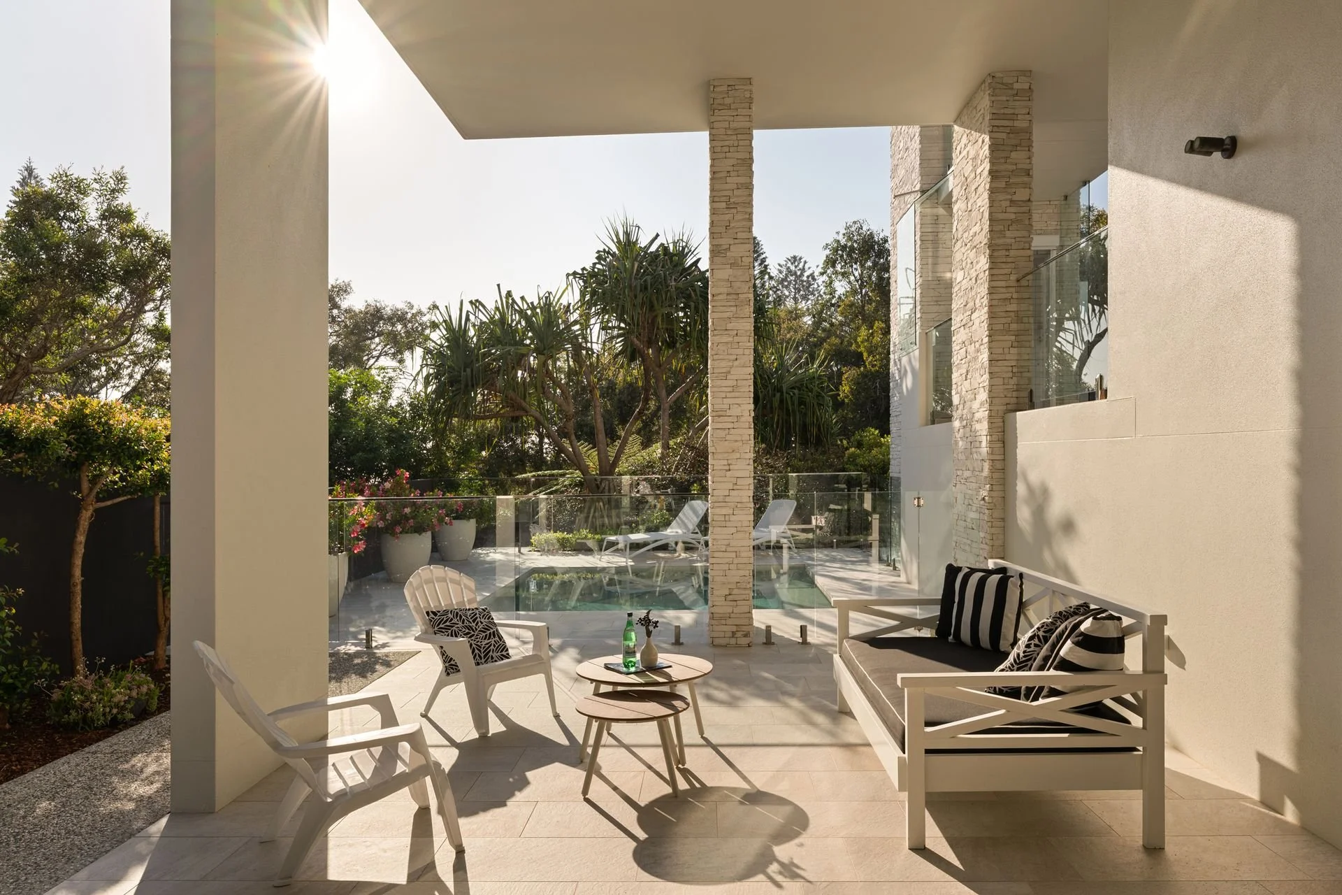 Modern patio with white furniture, black and white cushions, a small round table with a green bottle, and a background view of trees, potted plants, and a swimming pool, illuminated by sunlight.