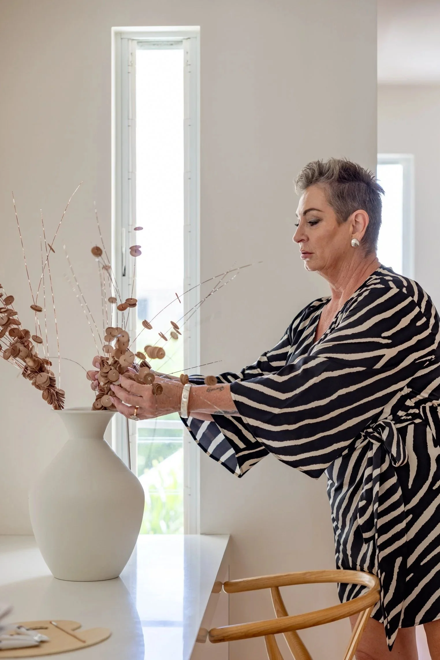 A woman with short gray hair and earrings, wearing a black and white striped robe, is arranging a flower arrangement in a white vase on a table near a window.