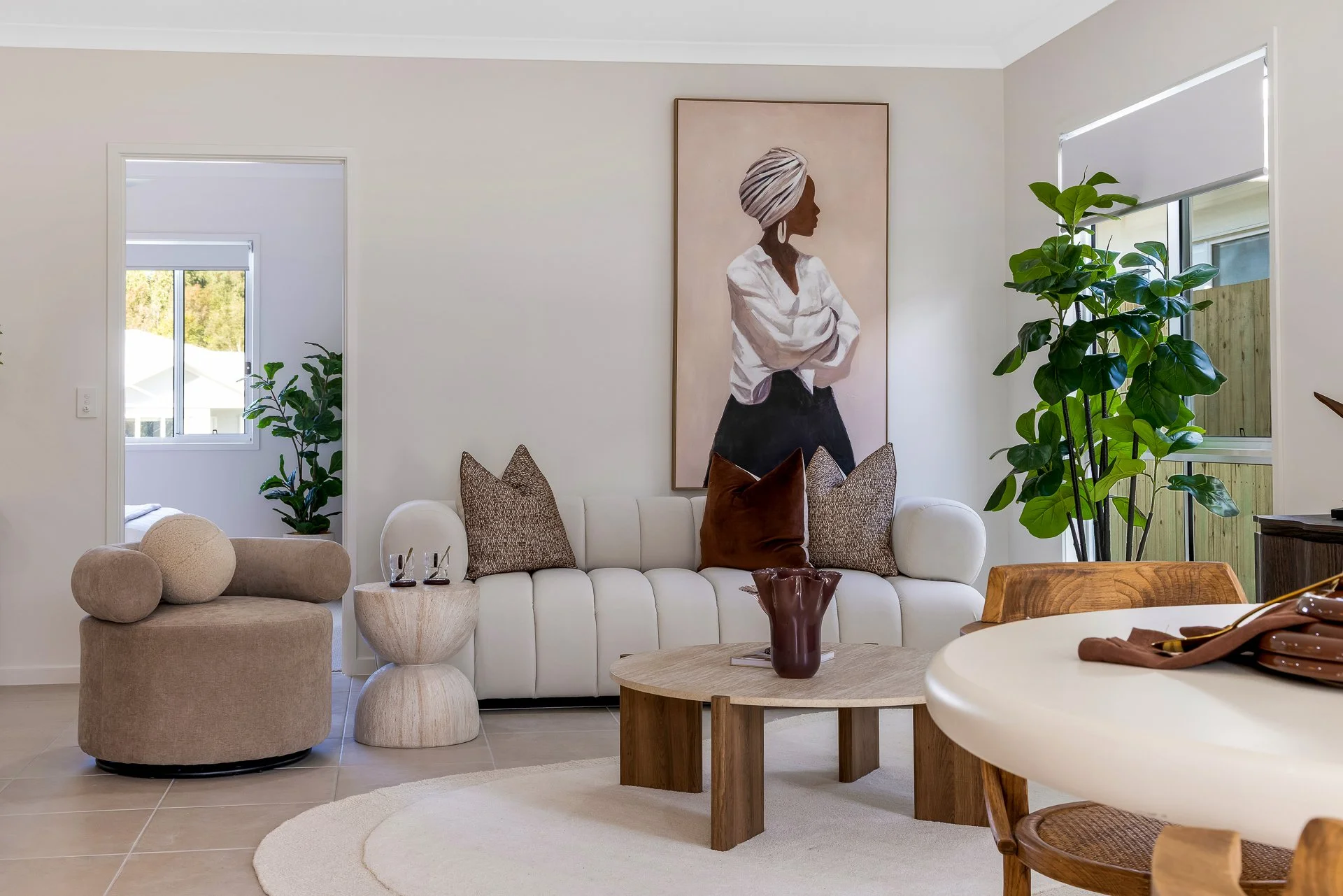 Living room with a white sofa, beige armchair, wooden coffee table, large potted plant, and artwork of a woman with a headwrap on the wall.