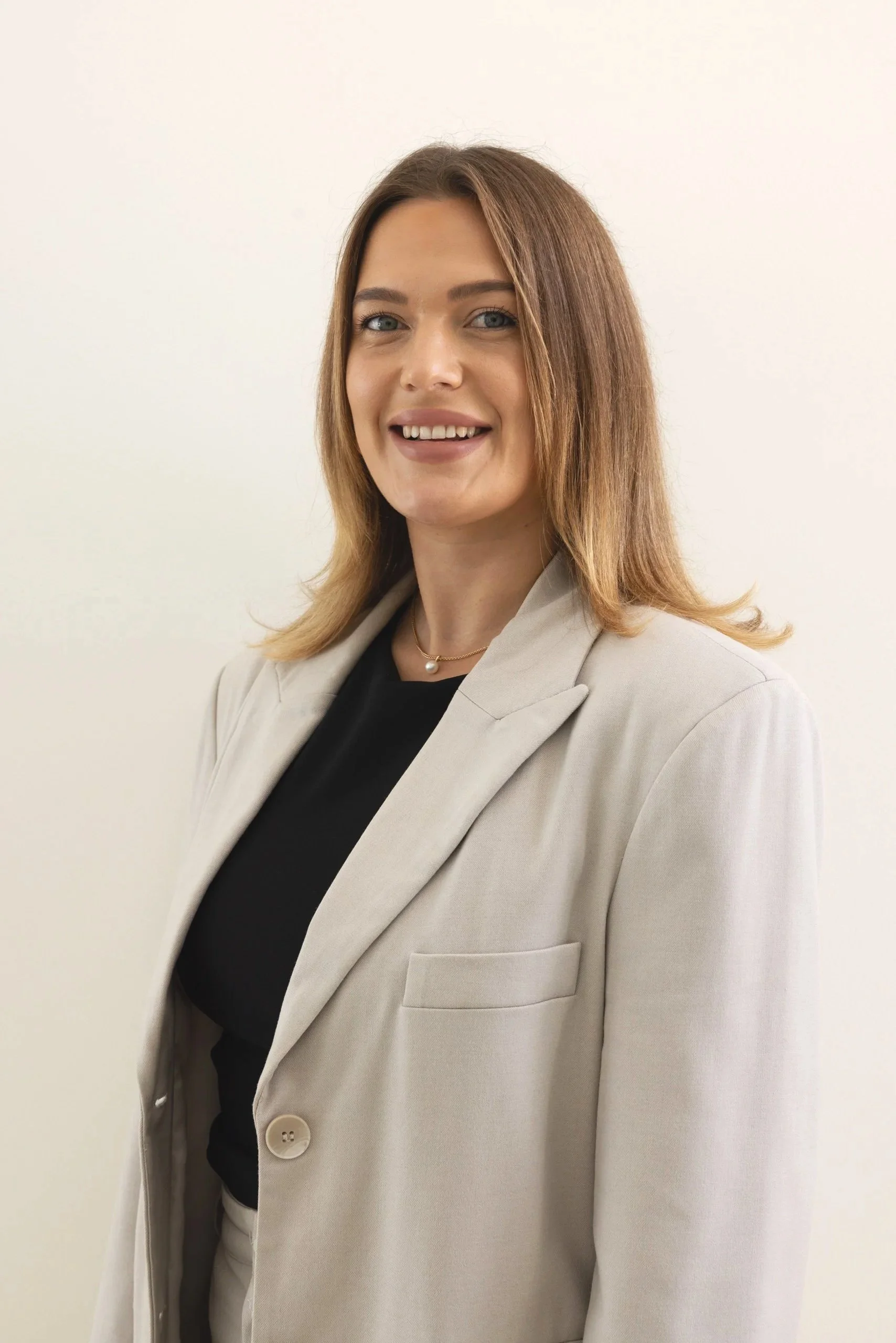 A woman with shoulder-length light brown hair and blue eyes, wearing a beige blazer over a black top, and a pearl necklace, standing against a plain off-white background.