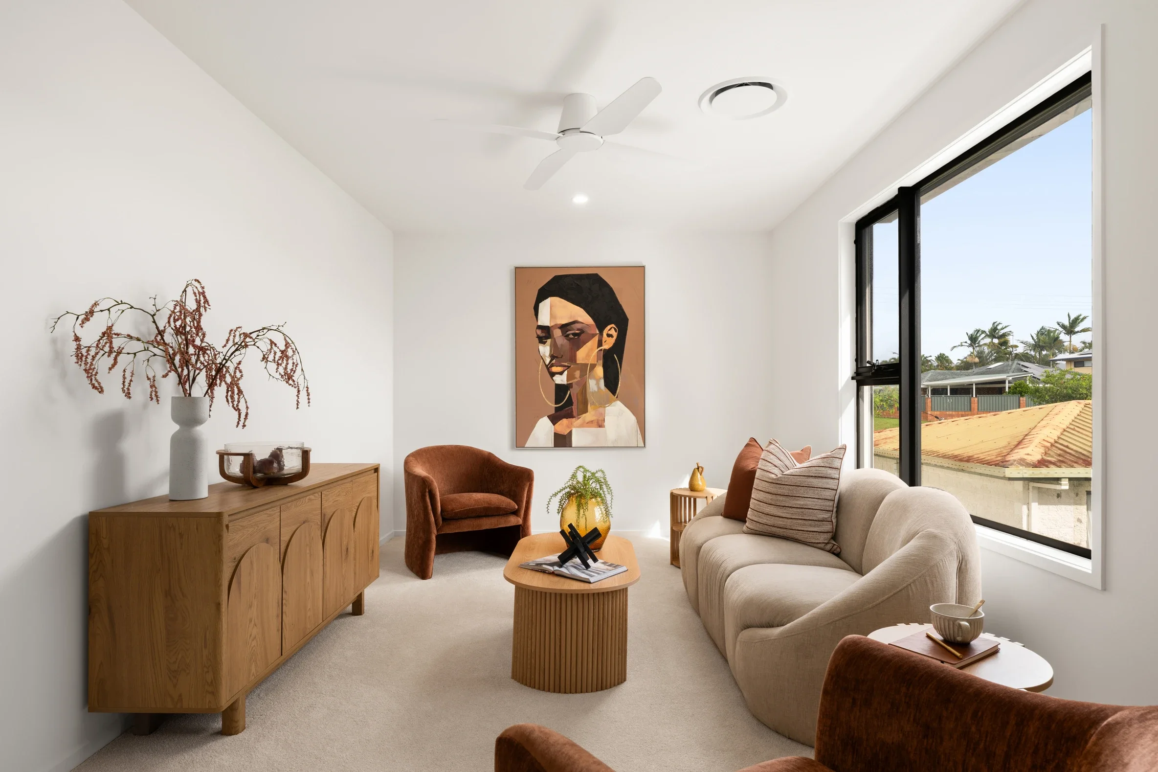 A modern living room with white walls, a beige sofa, and brown armchairs. Decor includes a large portrait of a woman, a plant, and a vase with dried branches on a wooden sideboard. Large window shows a view of houses and palm trees outside.