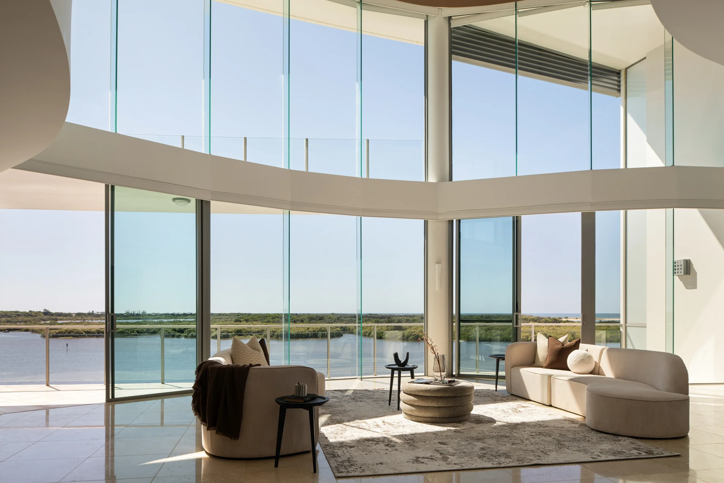 Modern living room with large glass windows overlooking a body of water and landscape, furnished with white sofas, a round textured coffee table, and side tables.