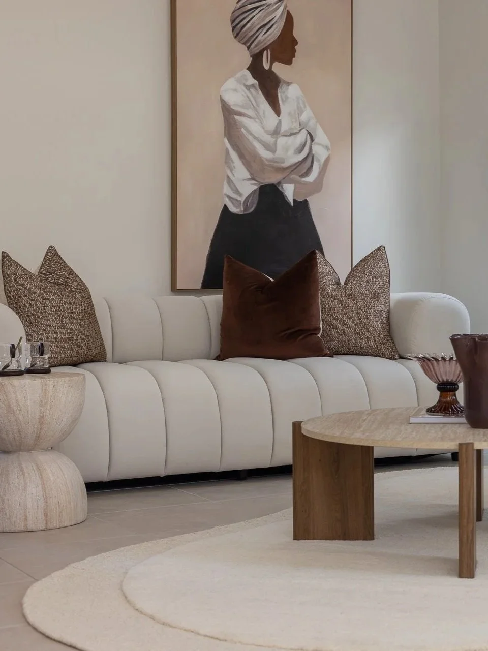 A modern living room with a white upholstered sofa, decorative pillows in brown and beige, a wooden coffee table with brown vases, and a large artwork of a woman in a white shirt and headscarf hanging on the wall.