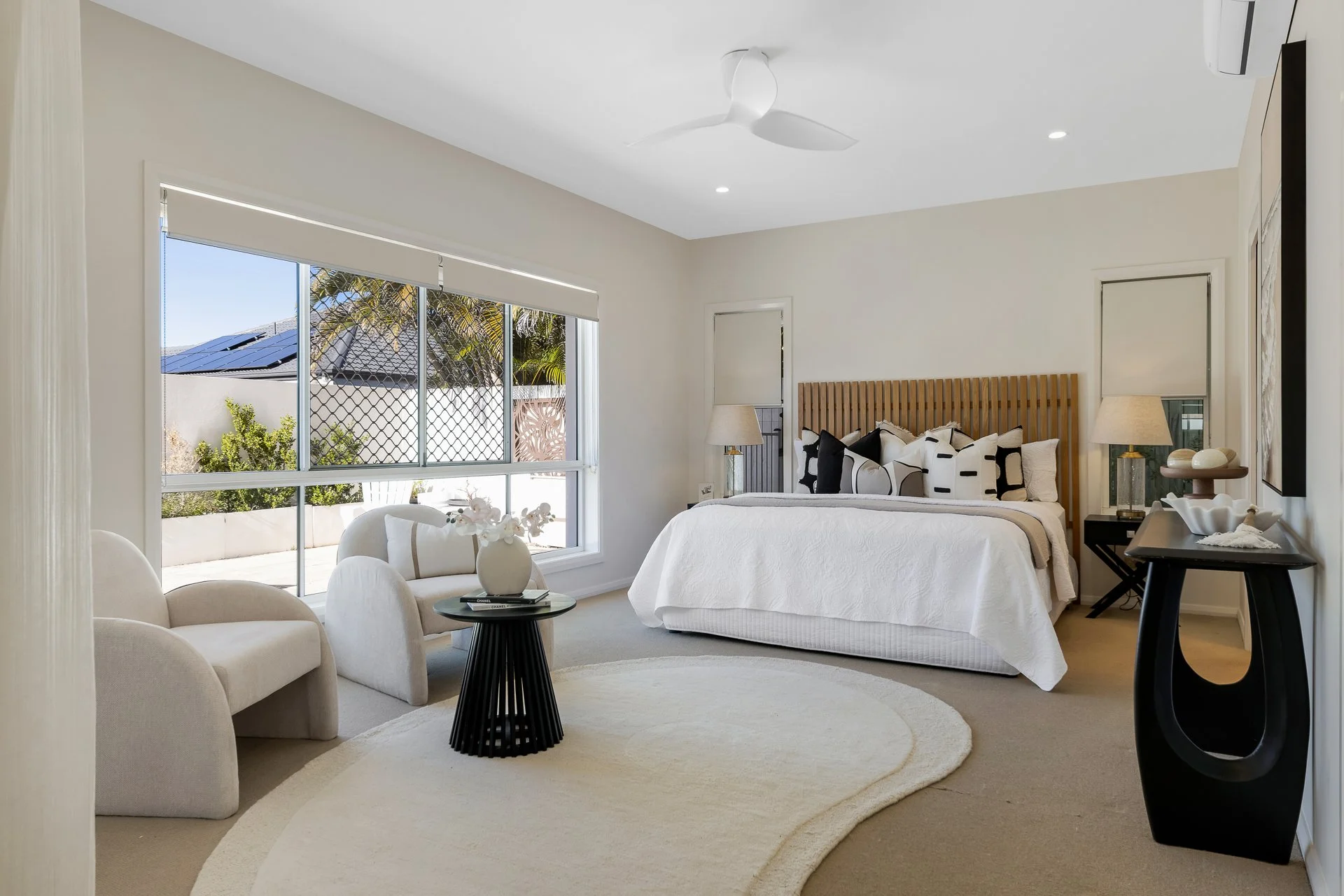 Modern bedroom with large window, white walls, tan carpet, bed with wooden headboard, black nightstand with white lamps, white chairs and round rug.