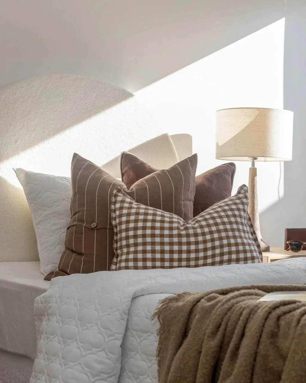 Sunlight streaming on a neatly made bed with a variety of decorative pillows, a bedside lamp, and a pair of sunglasses on a wooden nightstand.