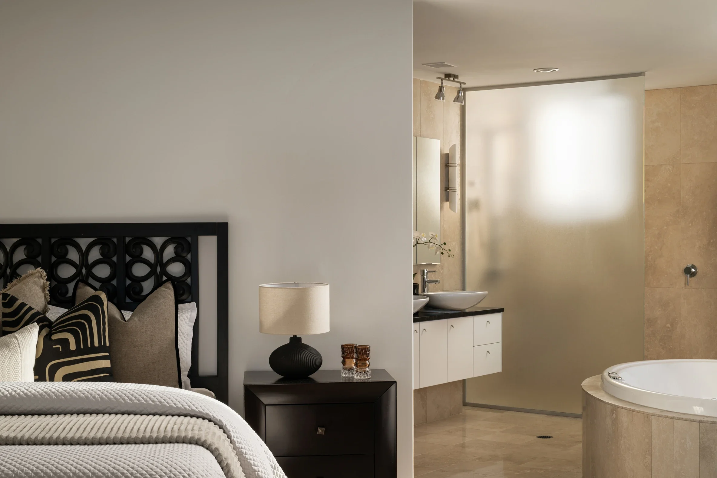 A bedroom with a black decorative headboard, beige and black throw pillows, a white textured bedspread, and a bedside table with a black lamp and decorative items. A large white wall separates the bedroom from a bathroom with sinks, a mirror, a frosted glass wall, and beige tiled floors.