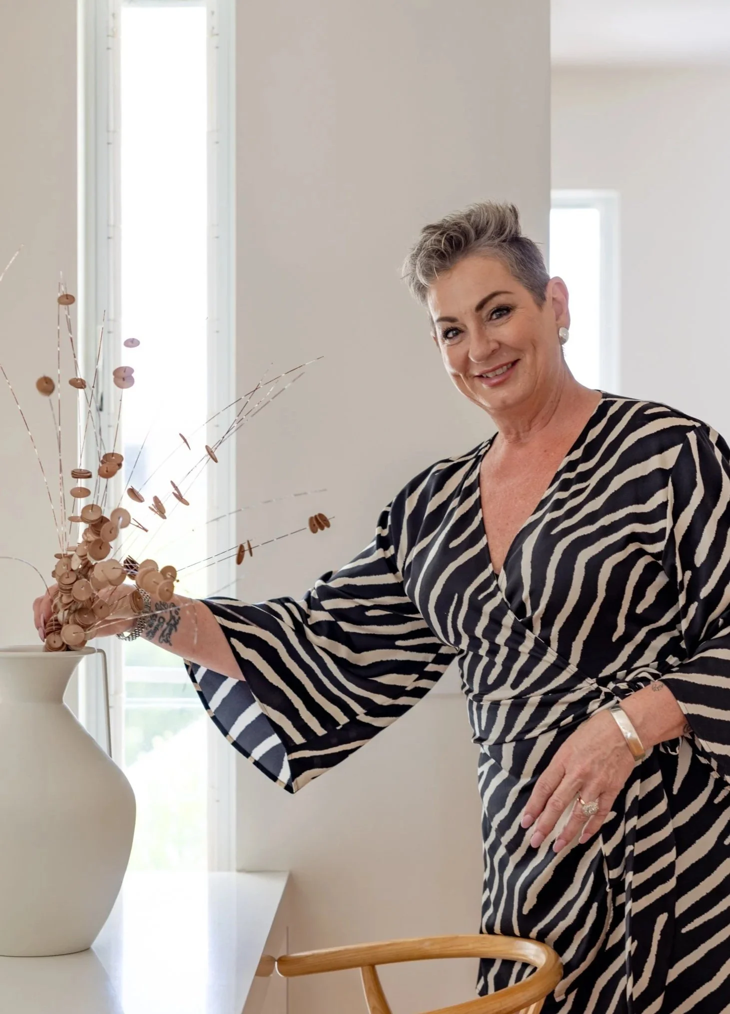 A woman with short, gray hair and a striped dress is smiling at a white ceramic vase filled with decorative branches. She is standing in a bright room with windows and a minimalist interior design.