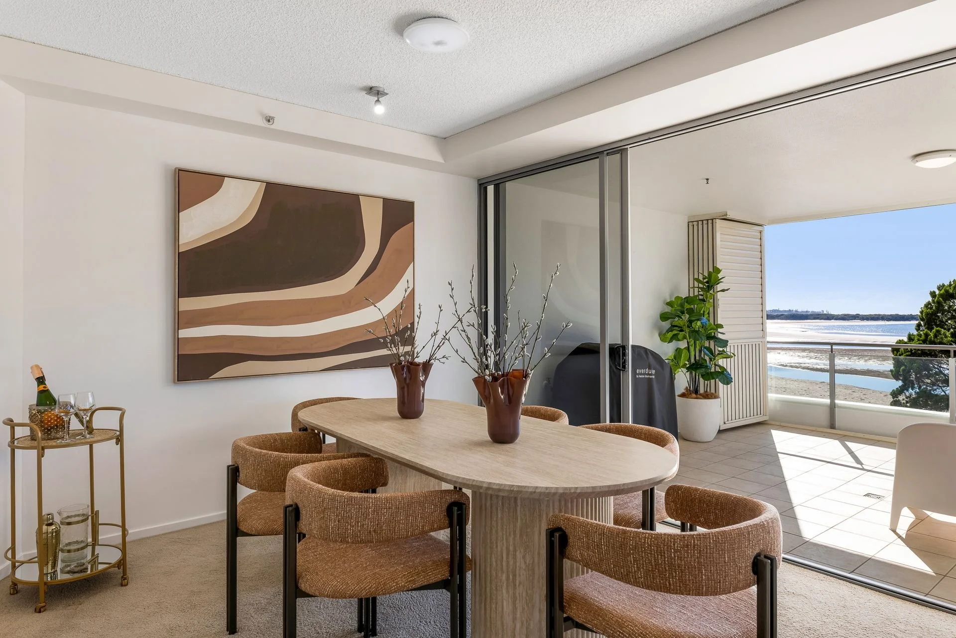 Dining room with a light wood table, six brown fabric chairs, two brown vases with decorative branches, abstract wall art, a glass wine cart with bottles and glasses, and sliding glass doors leading to a balcony with a view of the beach and ocean.