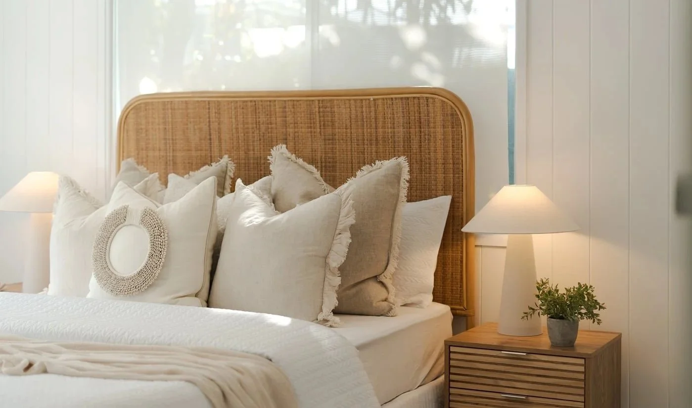 A cozy bedroom with a rattan headboard, white and beige throw pillows, a white bedspread, and a nightstand with a white lamp and a small potted plant.