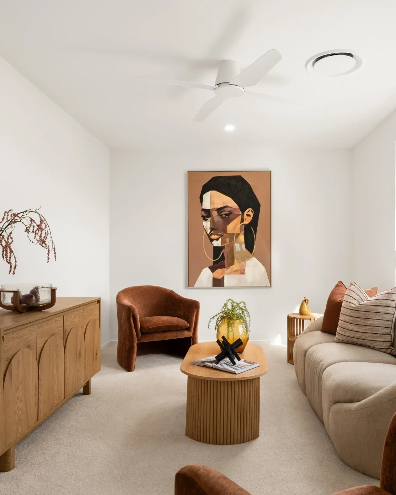 Living room with beige carpet, a beige sofa with striped and solid pillows, a brown armchair, a wooden sideboard, and a small wooden coffee table with decorative objects and a magazine. There is a framed portrait of a woman with dark hair on the wall, a potted plant on a yellow vase, some dried plant arrangements, and ceiling lights.