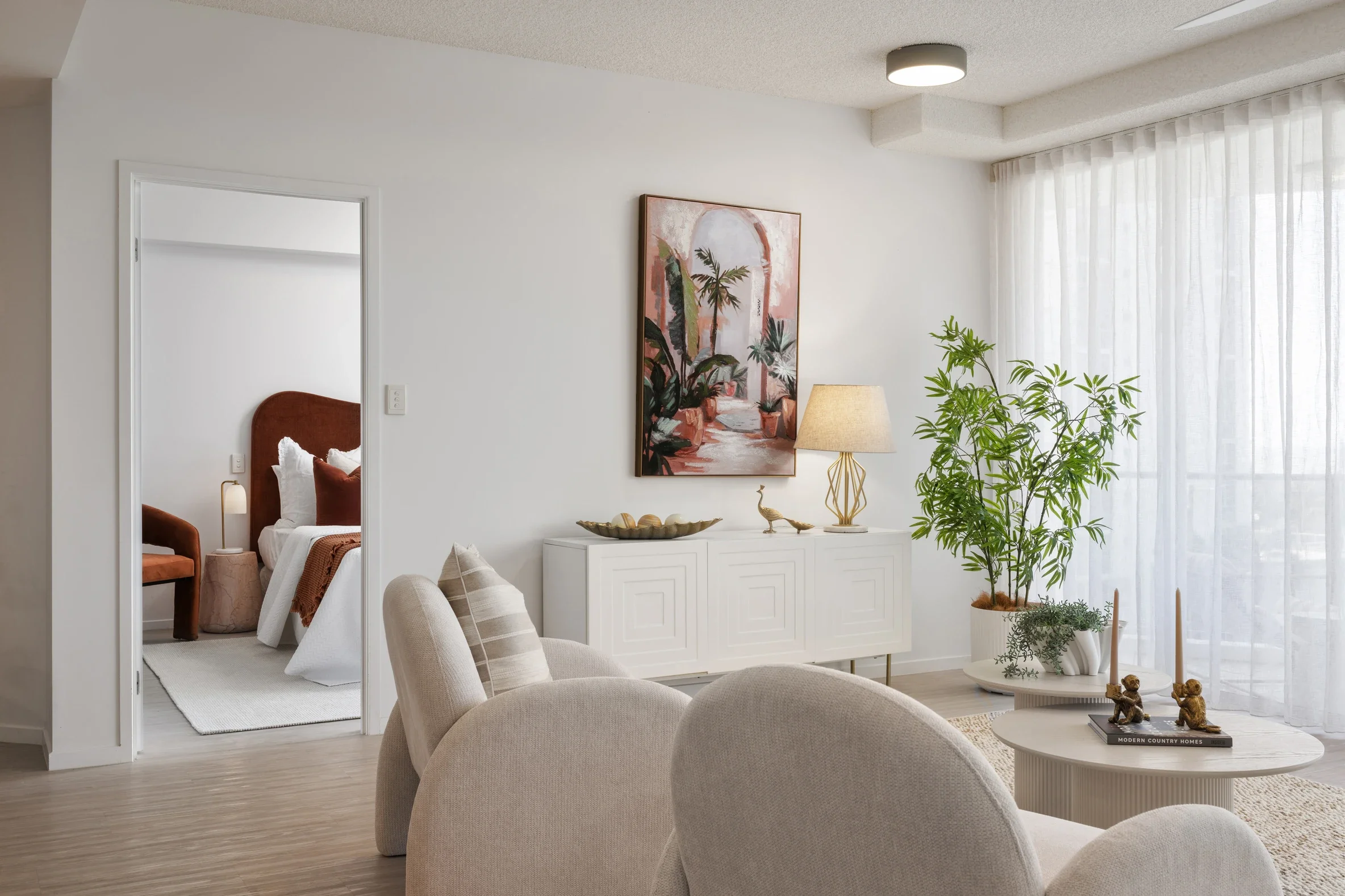 A modern living room with beige upholstered chairs, a white sideboard, and indoor plants near a large window with sheer white curtains. A large framed artwork of tropical plants decorates the wall.