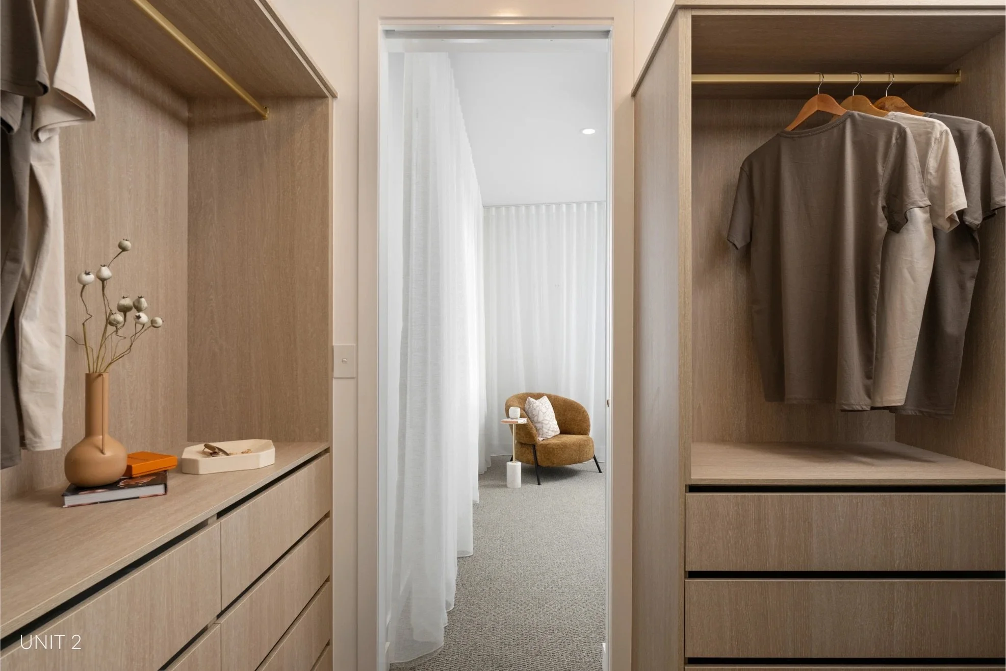 View of a walk-in closet with beige wooden shelves and hanging rods on both sides, some with hanging clothes. A decorative vase and some books are on a lower shelf on the left. At the end of the closet, through a doorway with a sheer white curtain, there is a small sitting area with a tan upholstered chair, a small white side table, and a cream-colored pillow. The room has neutral tones and a cozy atmosphere.