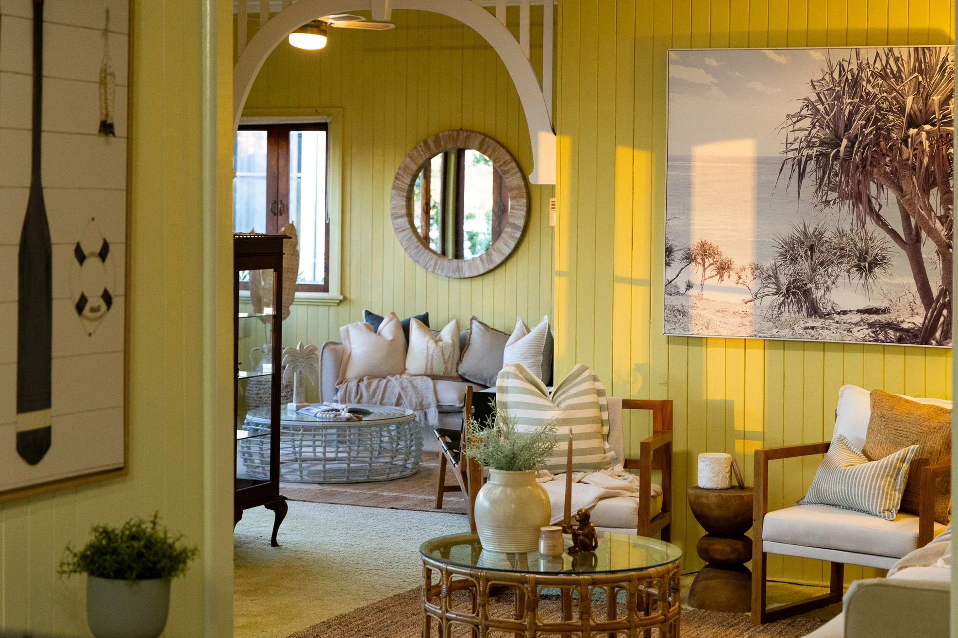 A cozy living room with a yellow-green paneled wall, featuring a large art print of a beach scene with trees. The room has a white sofa with pillows, a round wicker coffee table with decorative items, and a white vase with greenery. There is a small chair with striped fabric and a side table. The space is bright with natural light from a window.