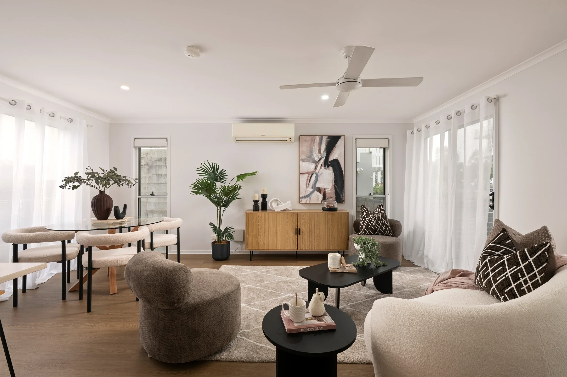 A living room with white walls, white curtains, a ceiling fan, and an art piece on the wall. There is furniture including a beige couch with patterned pillows, a black coffee table with books and decor, lounge chairs, a round dining table with beige chairs, and an assortment of plants and decorative items.
