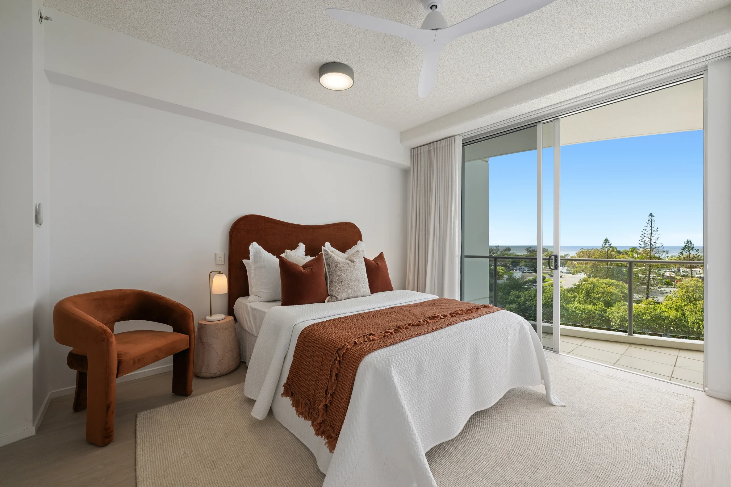Bedroom with a white bed, brown headboard, orange and white pillows, brown throw blanket, bedside lamp, brown velvet armchair, sliding glass door with balcony and ocean view, white curtains, ceiling fan, and modern ceiling light.