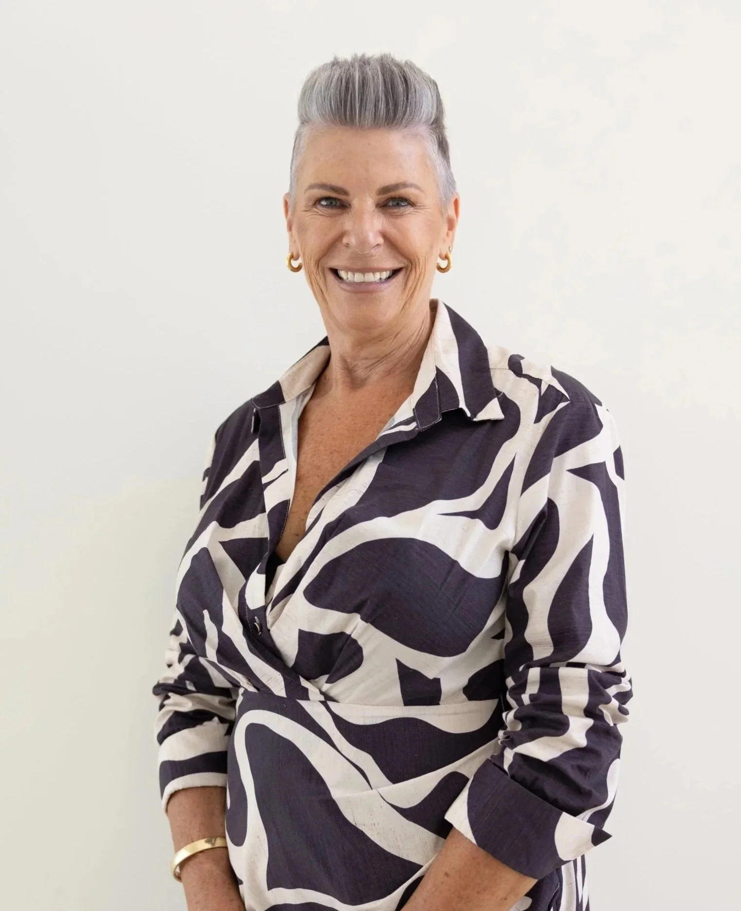 A woman with short gray hair smiling and wearing a black and white patterned shirt with gold earrings and a gold bracelet, standing against a plain white background.