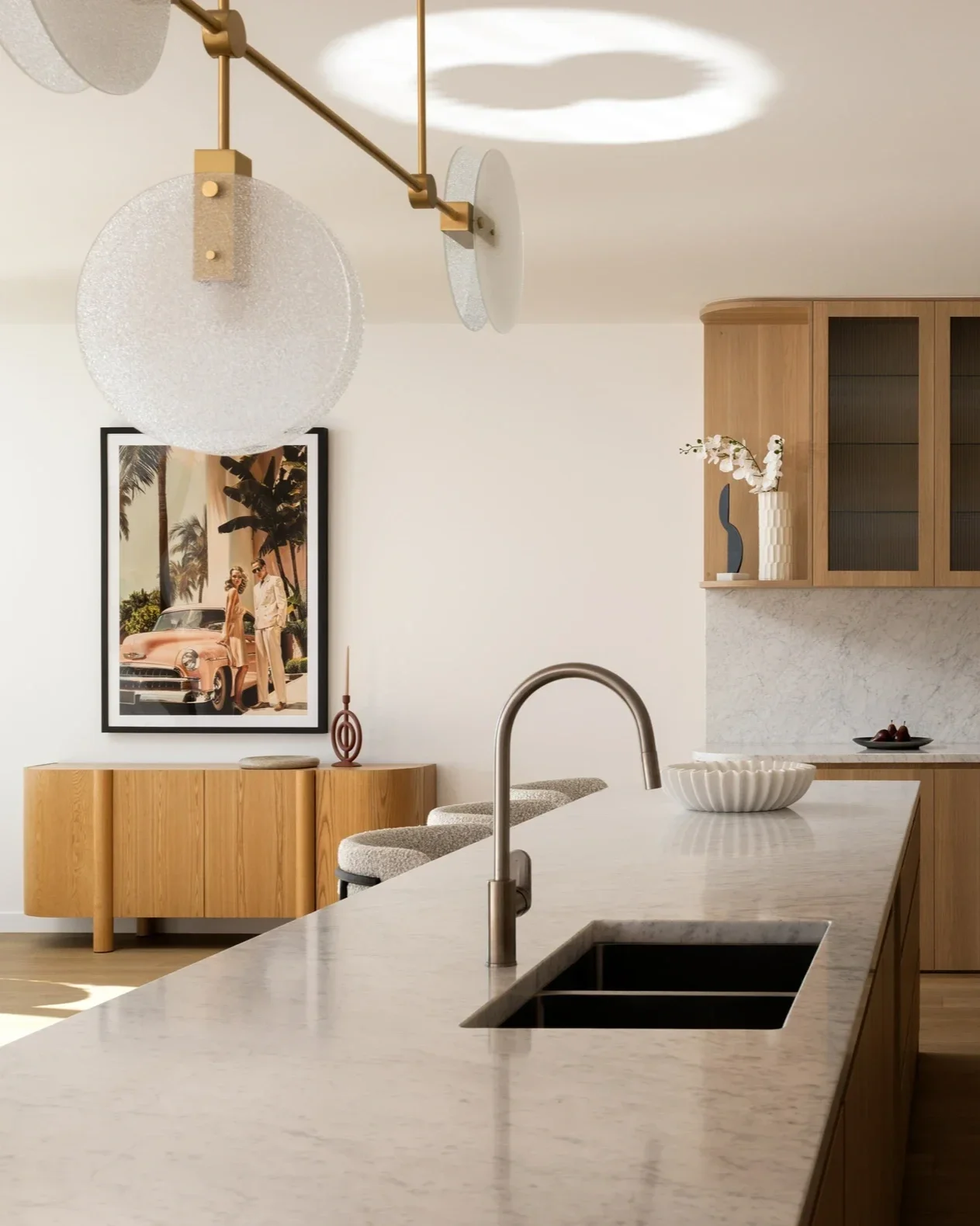 Modern kitchen with white marble island, wooden cabinets, a black sink, and minimalist decor including a large bowl of fruit. In the background, there is a wooden sideboard with a framed art print and a decorative vase with flowers. A contemporary chandelier with frosted glass globes hangs from the ceiling.