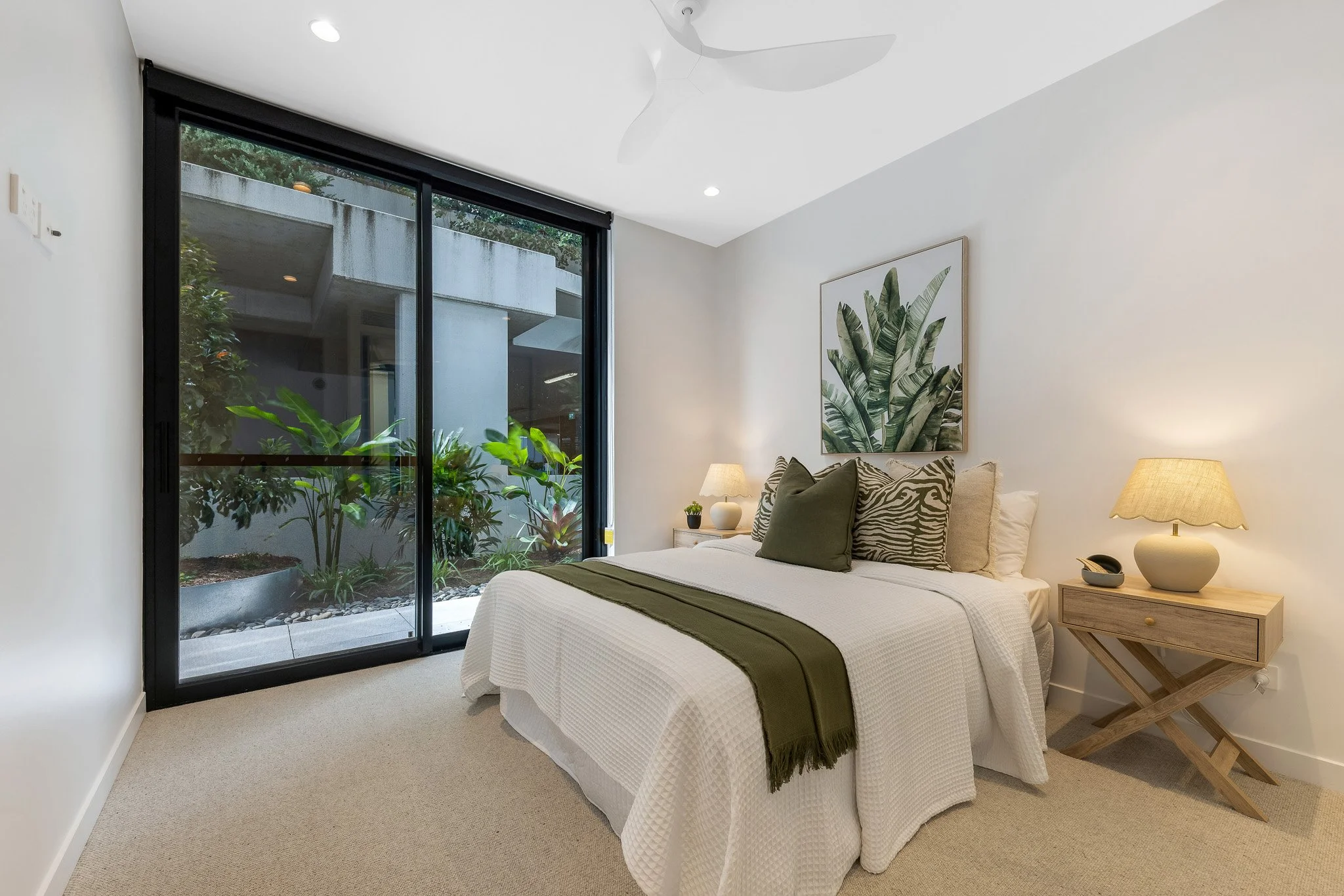 A bedroom with a double bed covered in a white quilt, green and zebra-print pillows, and a green throw. A wooden bedside table with a beige lamp, small plant, and decorative bowl is next to the bed. A large framed botanical print hangs above the bed. There is a large sliding glass door leading outside, with a view of greenery and an outdoor patio. The room has beige carpet and white walls, with a white ceiling fan.
